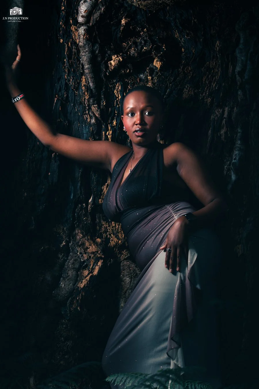 A woman in a dark dress with a gradient from black to beige, standing against a textured dark rock wall, striking a pose with one arm raised and the other resting on her waist.