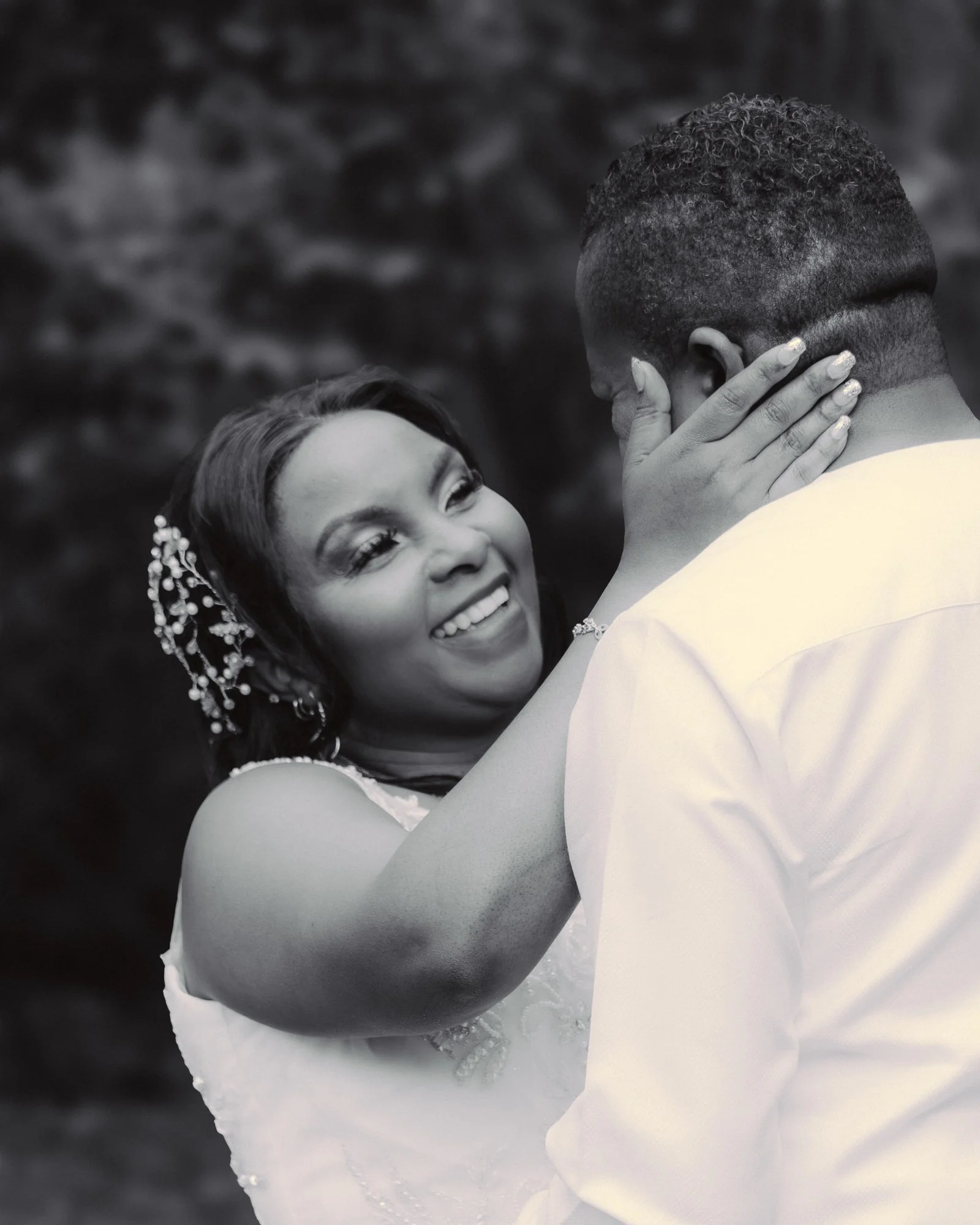 A joyful black couple sharing an intimate moment, with the woman smiling and touching the man's face, both dressed in wedding attire against a blurred outdoor background.