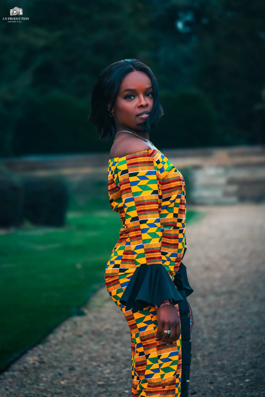 A woman with short black hair wearing a colorful, traditional African dashiki with geometric patterns, standing outdoors on a gravel path with greenery in the background.