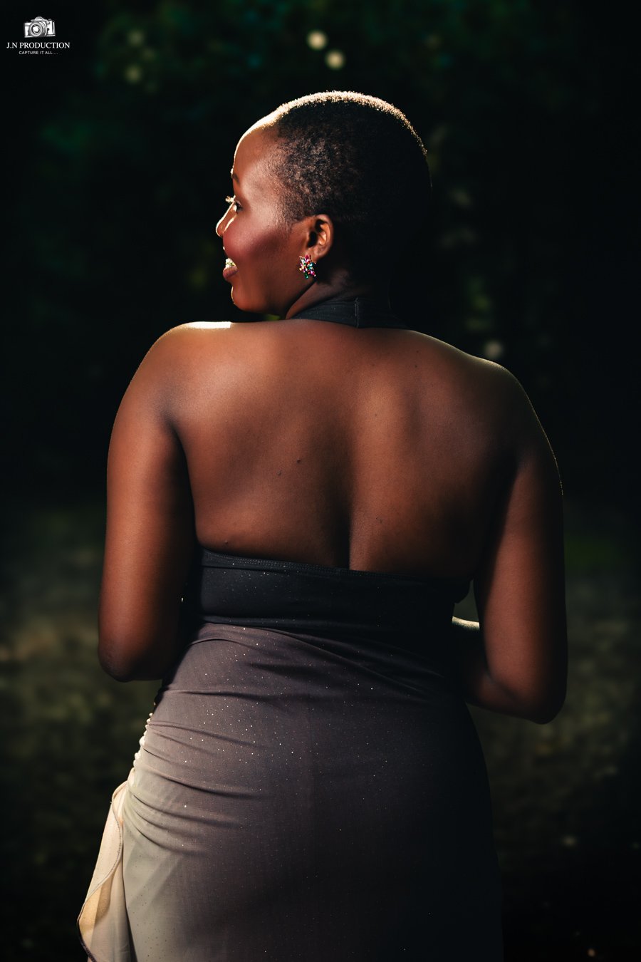 A woman with short, natural hair, wearing colorful earrings, facing sideways with her back and profile visible, illuminated by soft lighting, outdoors with a blurred dark green background.