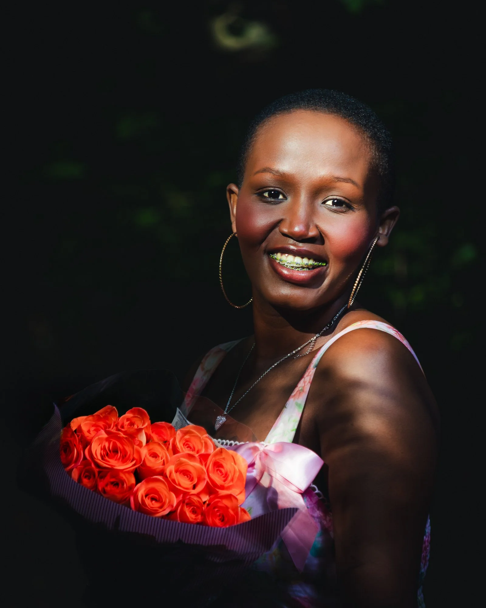 A woman smiling while holding a bouquet of orange roses, wearing a colorful dress, hoop earrings, a necklace, and braces, against a dark background with some green foliage.