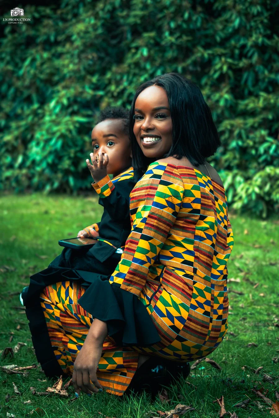 A woman and a young boy sitting on the grass in front of a green bush. The woman is smiling and wearing a colorful patterned traditional African dress. The boy is holding a smartphone and has his hand near his mouth.