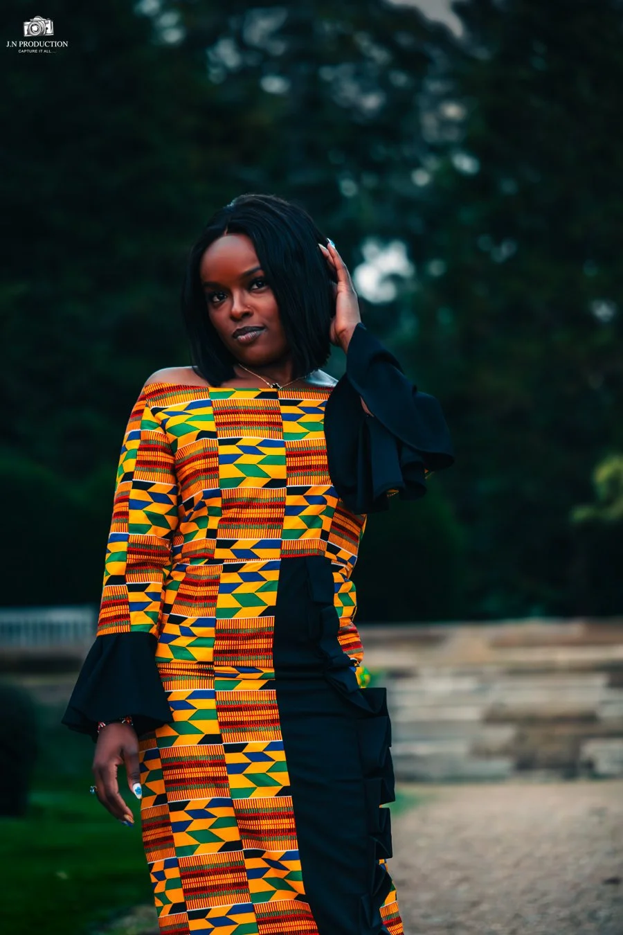 A woman in a colorful African print dress with geometric patterns in yellow, green, red, and blue, standing outdoors with trees in the background.