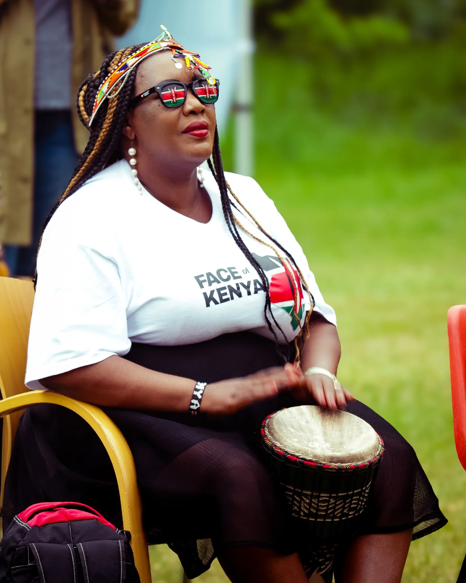 Woman wearing sunglasses and beads, playing a drum outdoors during a cultural event, with a white T-shirt that says 'Face of Kenya' and the Kenyan flag on it.