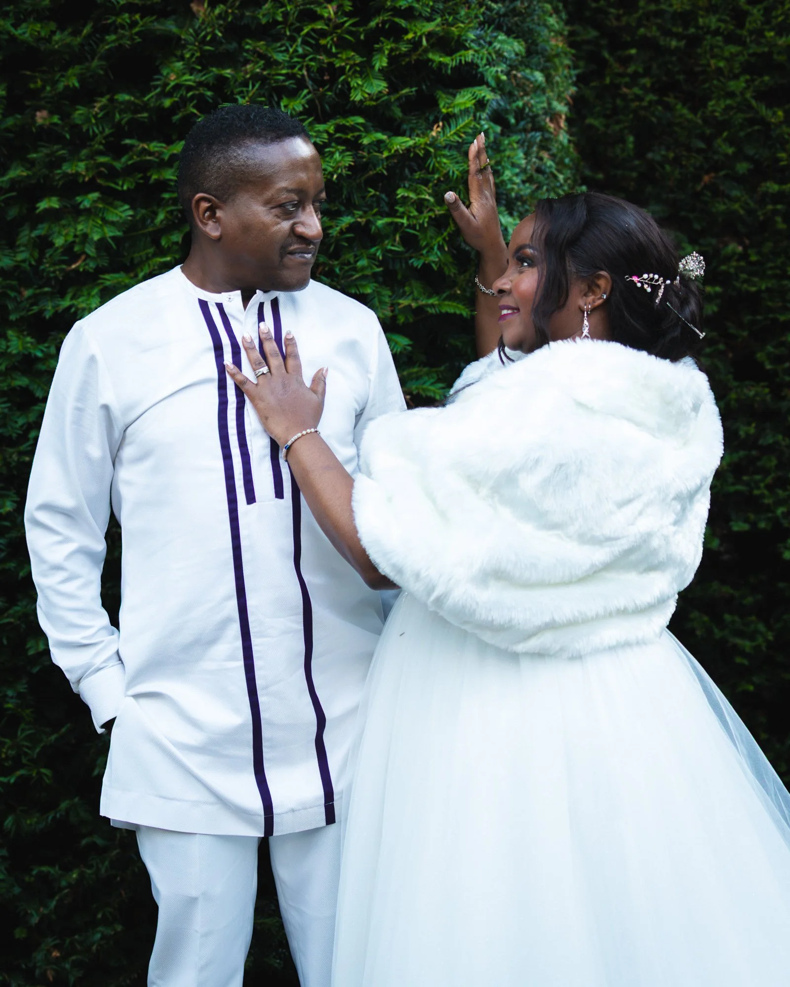 A man and woman dressed in formal attire facing each other, with the man in a white traditional outfit with purple accents and the woman in a white gown with a fur shawl, standing outdoors against a background of green foliage.