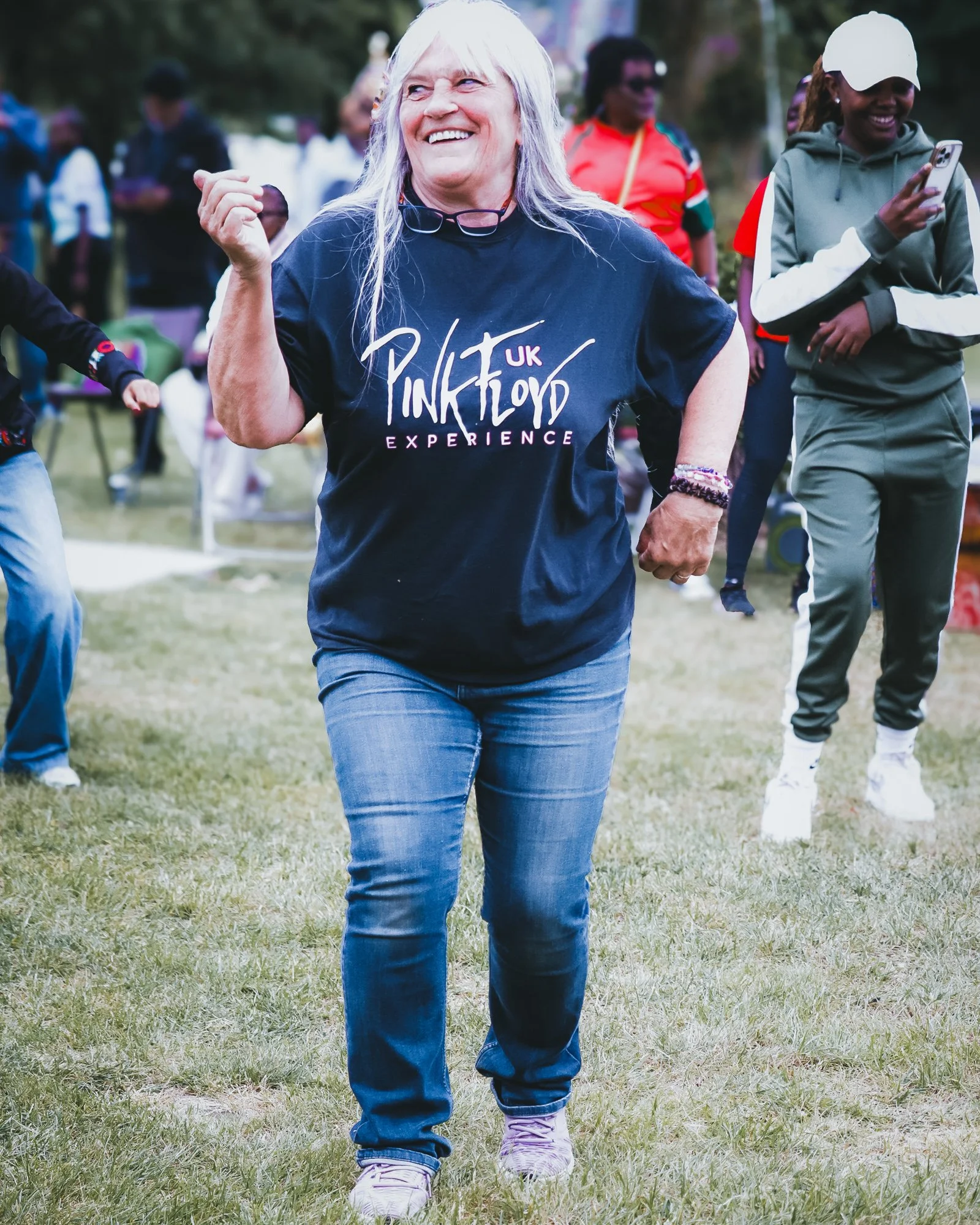 A smiling woman with long white hair participating in an outdoor Pink Floyd UK Experience event, surrounded by other people, some taking photos and others walking on a grassy field.