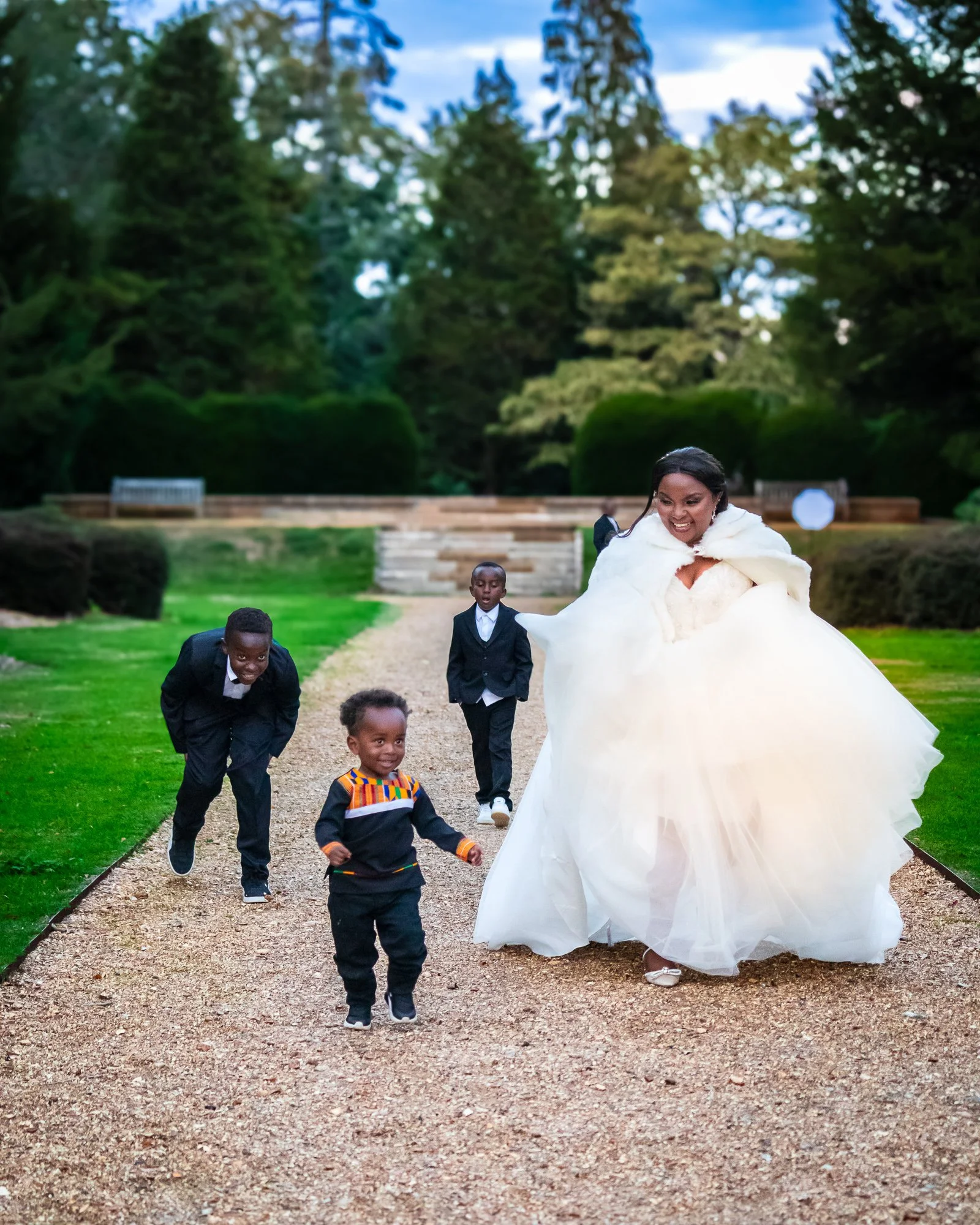 A woman in a white wedding dress running on a gravel path with three children, two boys and one girl, all dressed in formal attire, in a lush outdoor garden setting.