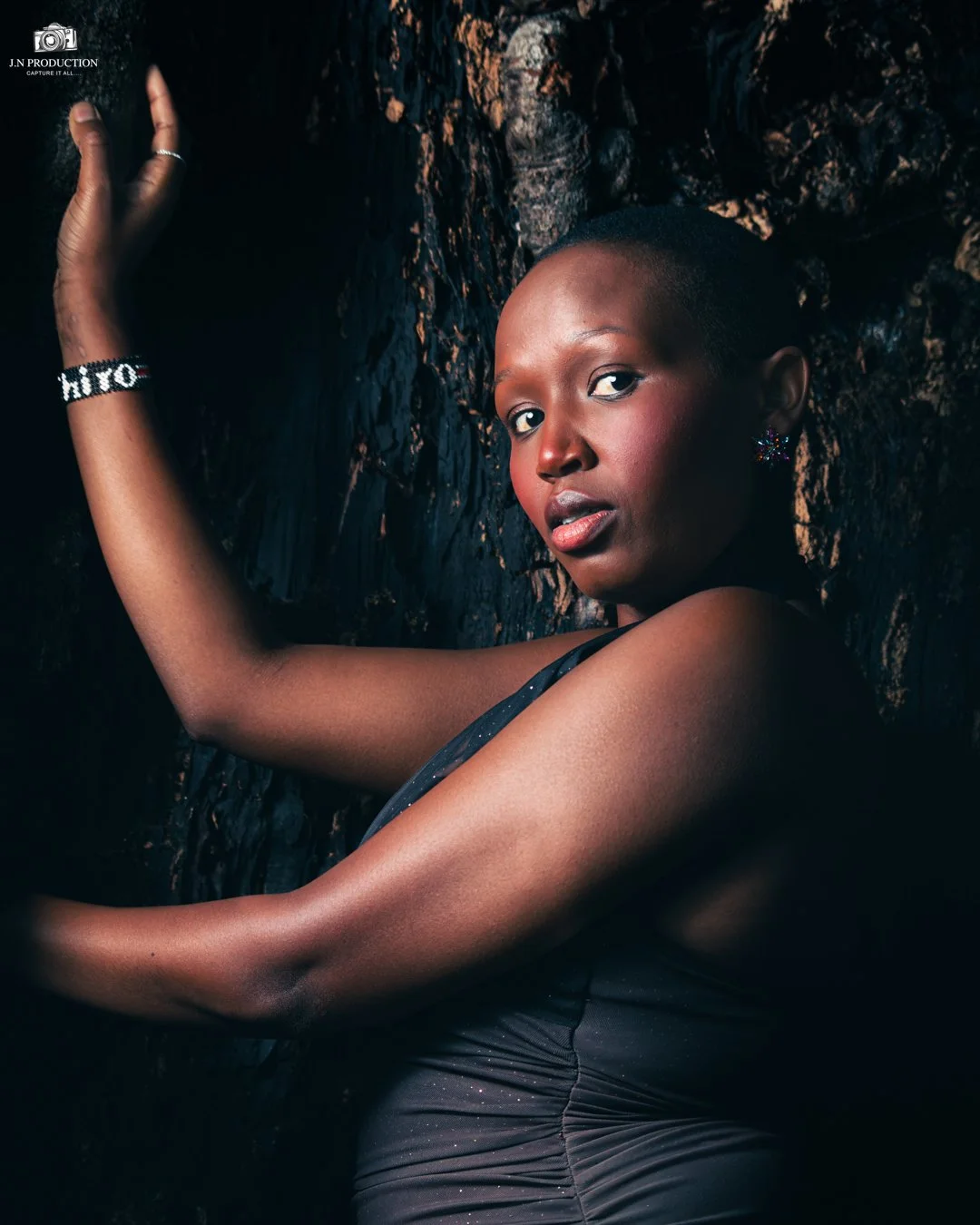 A portrait of a young woman with short hair, striking a pose against a dark, textured background. She is wearing a black sleeveless top and jewelry, including a bracelet with the name 'Hiro' and sparkly earrings. She has a confident expression, with 