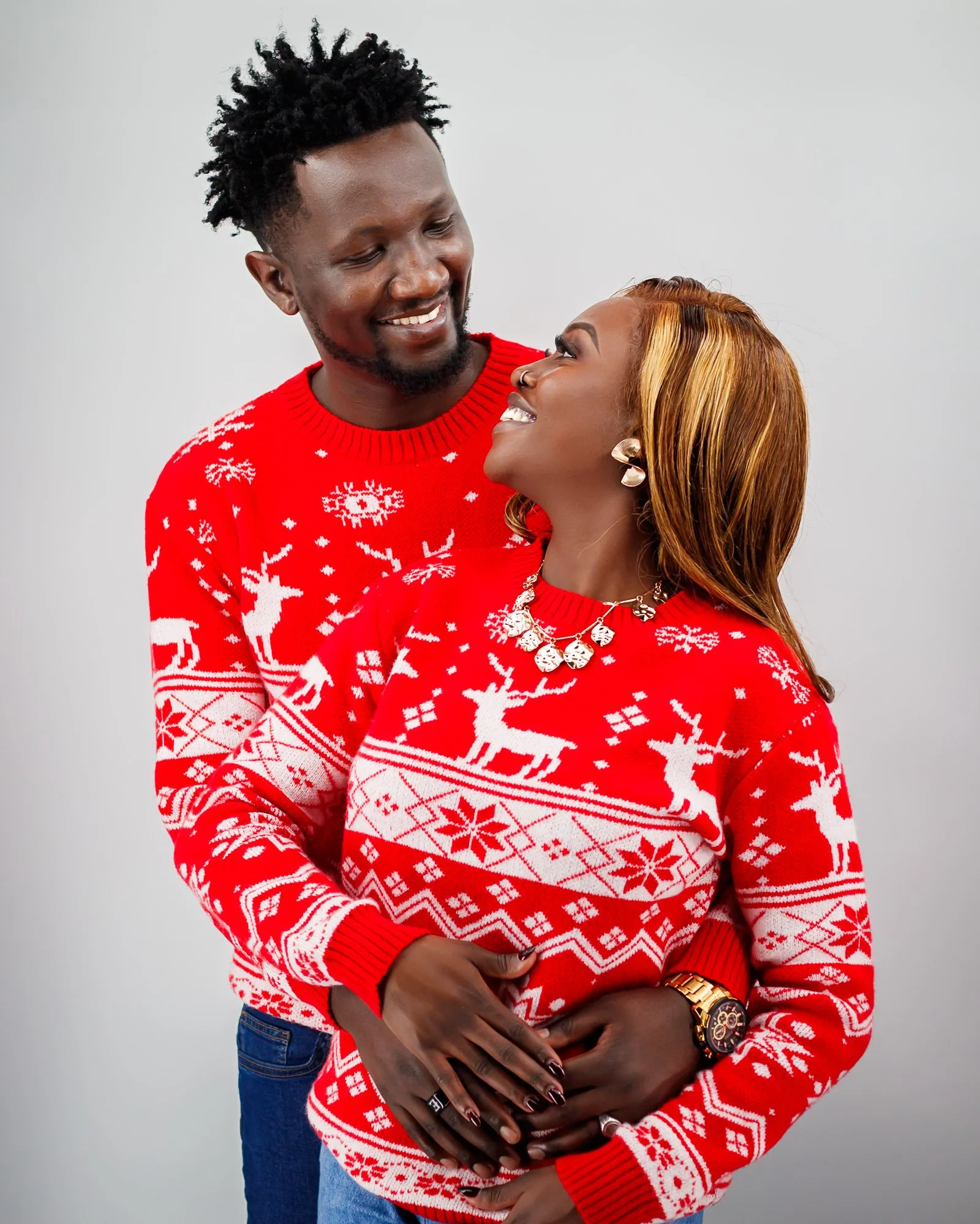 A smiling couple wearing matching red Christmas sweaters with white reindeer and snowflake patterns, standing against a plain white background, sharing a joyful moment.