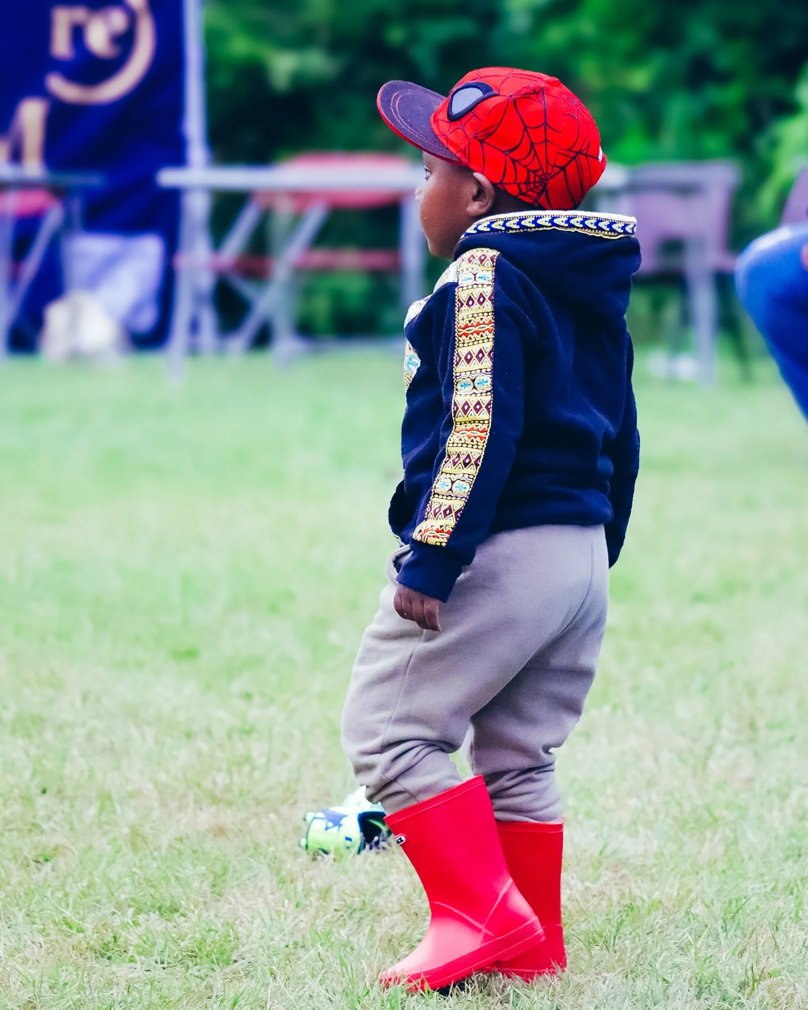 A young child stands on a grassy field wearing a red Spider-Man cap, a navy hoodie with colorful embroidery on the sleeves, beige pants, and bright red rain boots. There are blurred tents and people in the background.