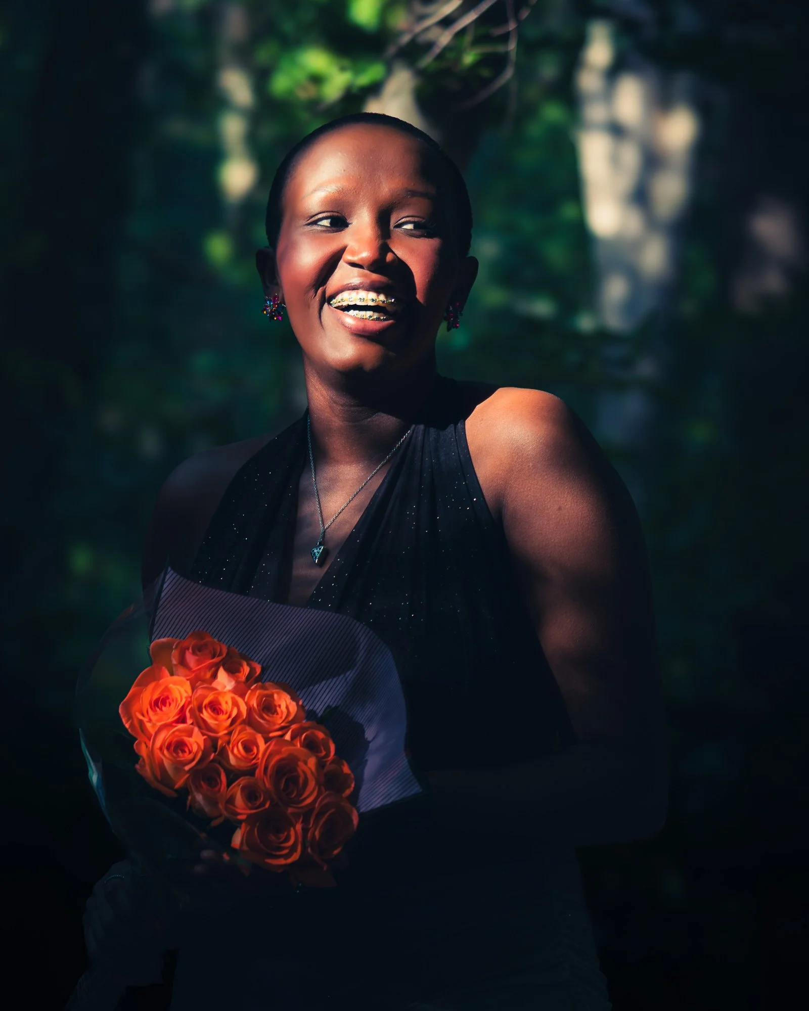 A woman with dark skin smiling, holding a bouquet of orange roses, wearing a black dress, jewelry, and braces, outdoors with dark background and sunlight on her face.