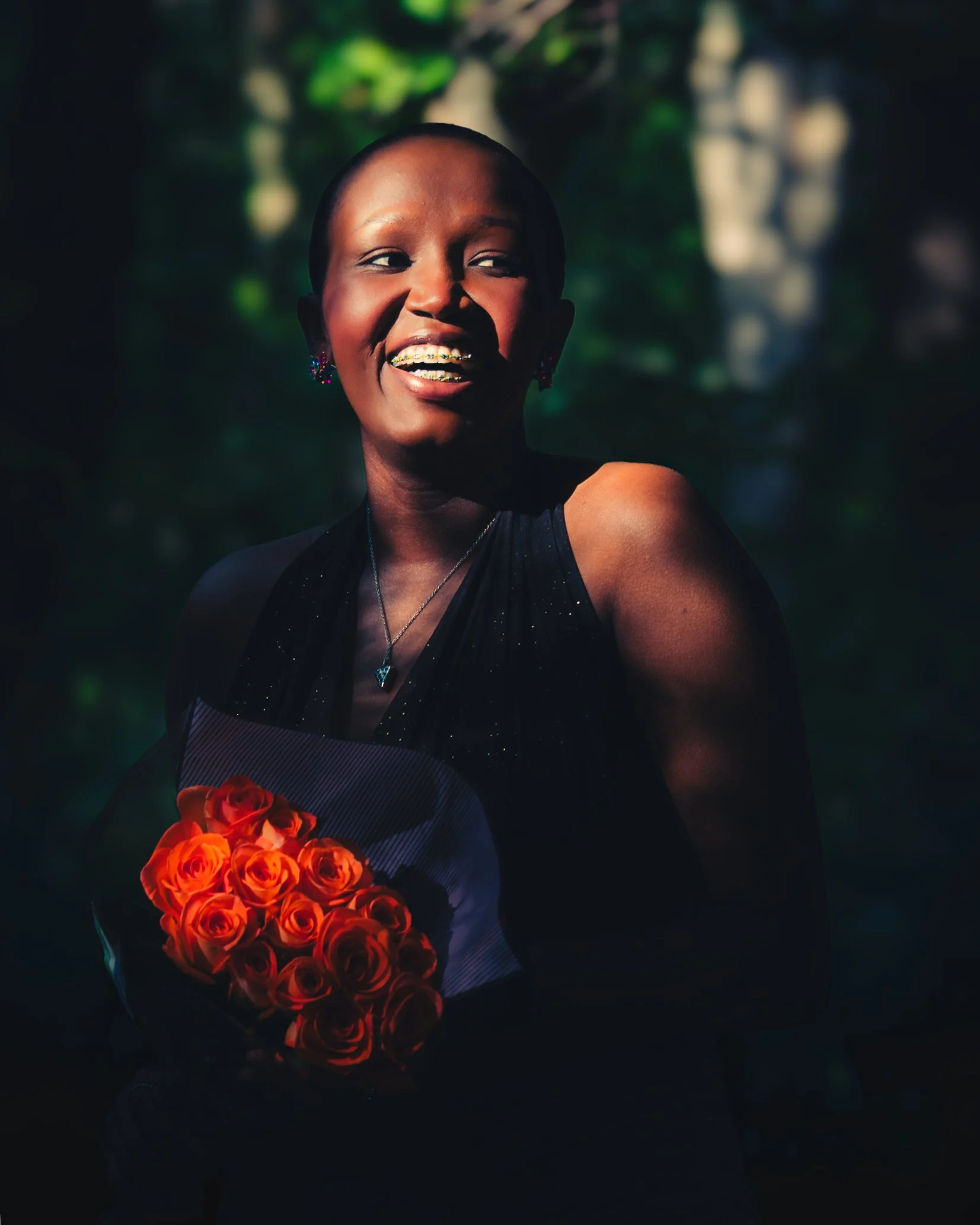 A woman smiling and holding a bouquet of orange roses, outdoors with a dark, blurred background.