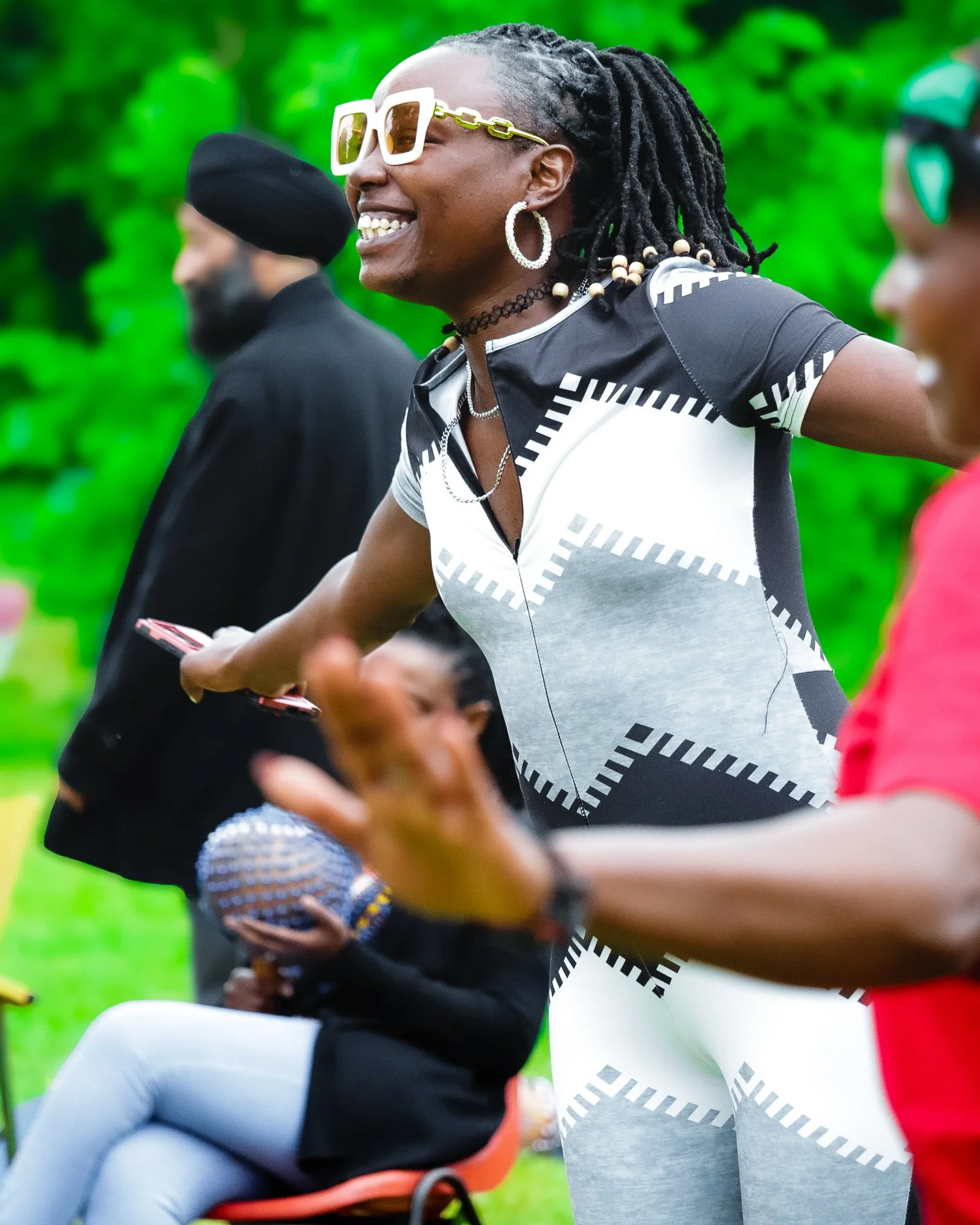 A woman with dreadlocks, wearing white sunglasses, hoop earrings, and a patterned black and white dress, smiling and engaging with others outdoors.
