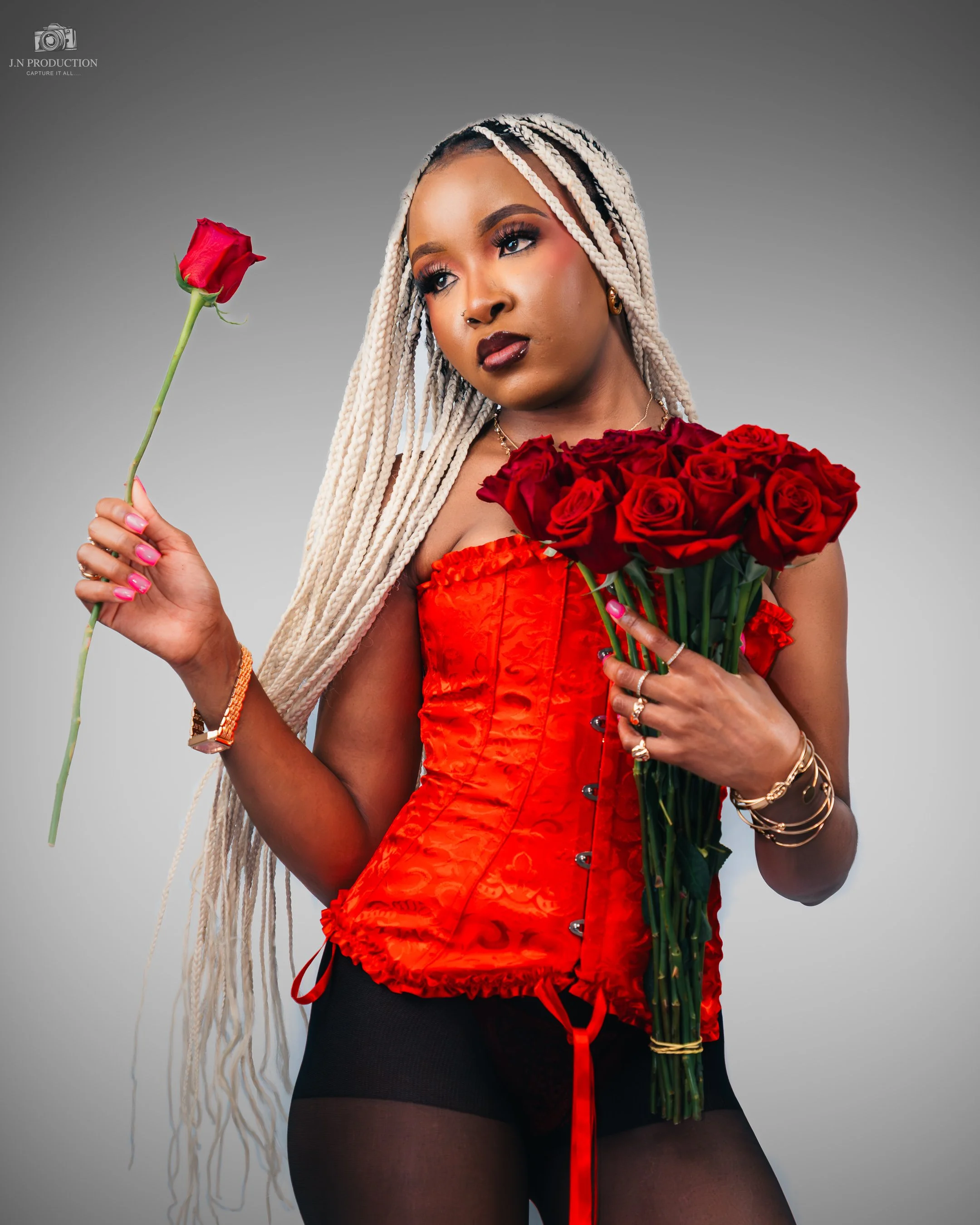A woman with long blond braids wearing a red corset top and black stockings holds a bouquet of red roses and a single rose in her hand, standing against a gray background.