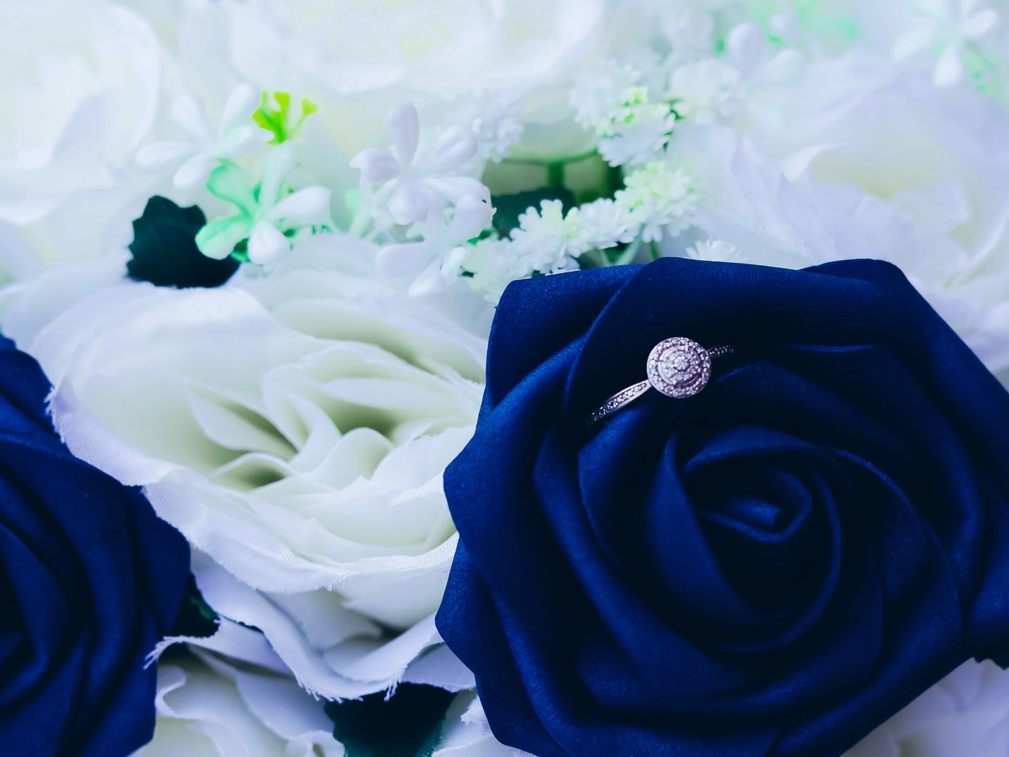 A close-up of a jewelry ring placed on a navy blue artificial rose surrounded by white flowers and green accents.