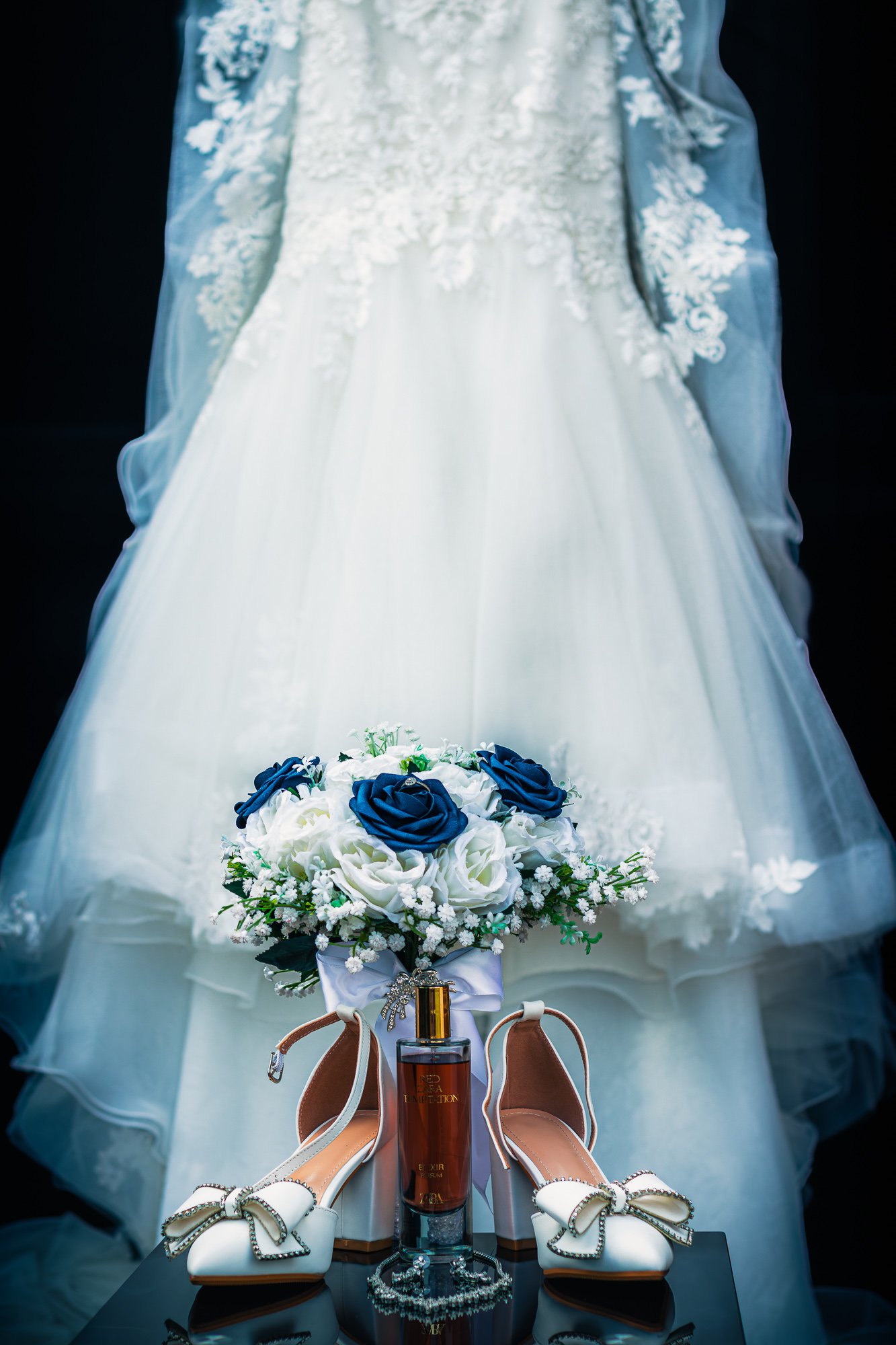 Details of a white wedding dress, a bouquet with white and blue roses, a pair of white heels with bows, perfume bottle, and jewelry displayed on a black table.