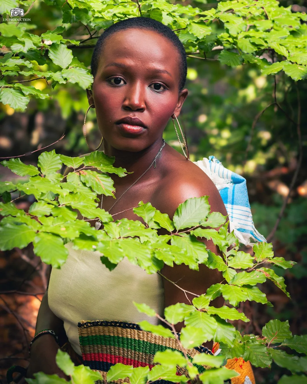 A woman with dark skin and short hair stands amidst green leaves, looking directly at the camera. She is wearing earrings, a necklace, and a strapless top with colorful patterns.