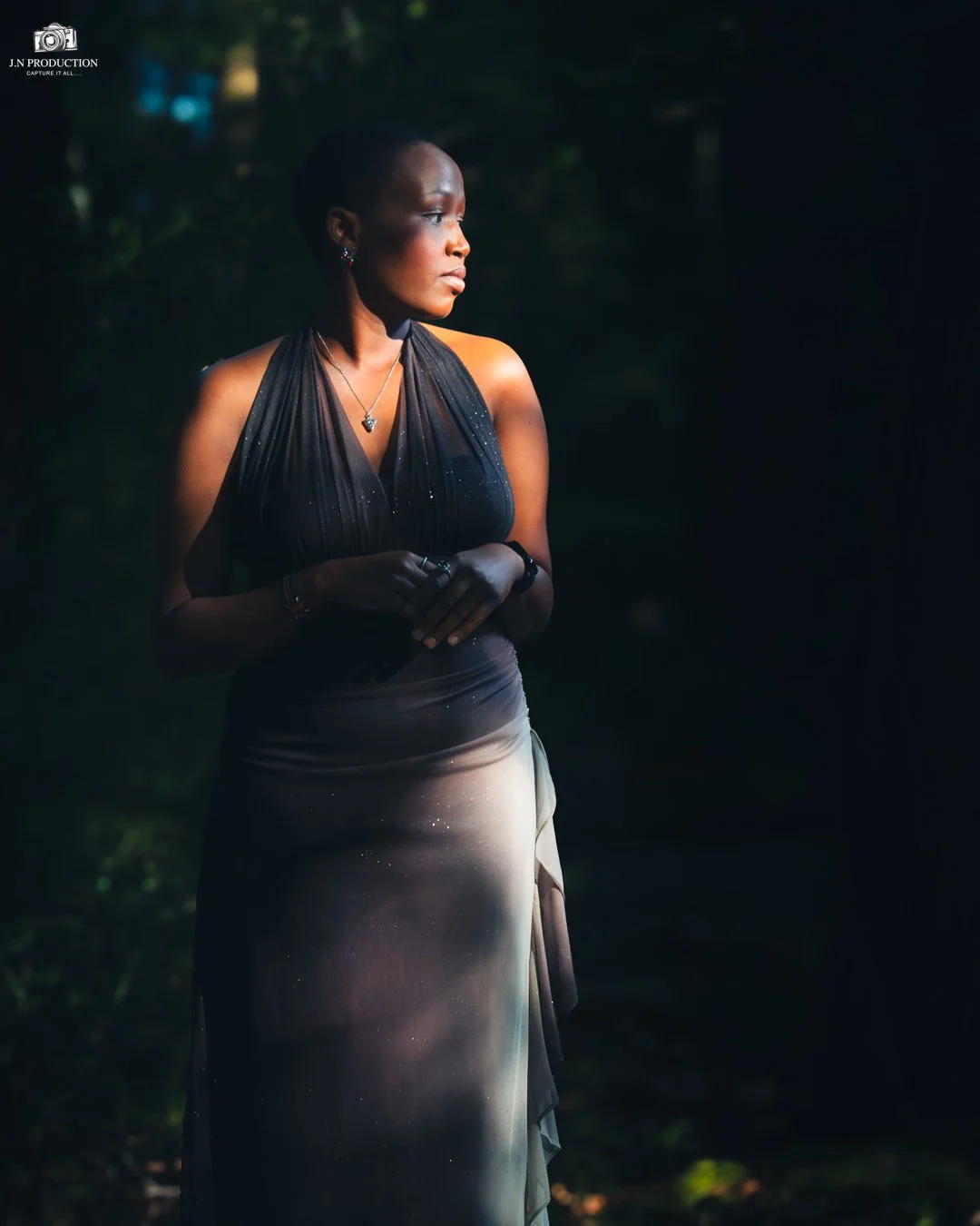 A woman with short hair wearing a black sleeveless top and a long, gradient skirt stands outdoors in a dark, forested area. She is looking to her left with her hands clasped together in front of her.