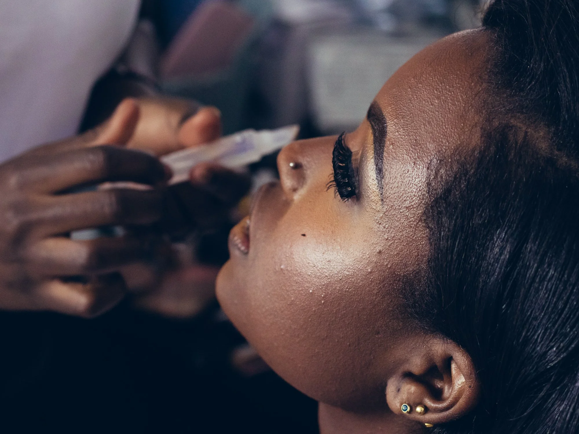 A woman with long black hair and gold earrings has makeup applied by a makeup artist. The focus is on her face as the makeup artist works on her lips.