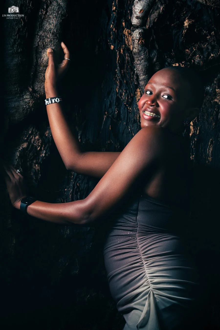 A woman with a shaved head smiling and hugging a tree trunk in a dark setting.
