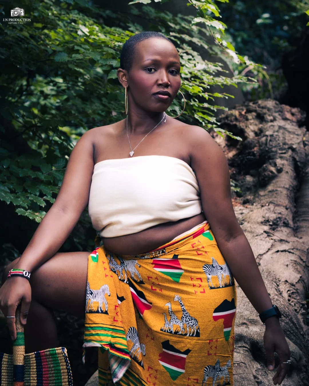 A woman with short hair and hoop earrings sitting outdoors among green foliage and rocks, wearing a strapless cream top and a colorful wrap skirt with zebra and animal patterns.