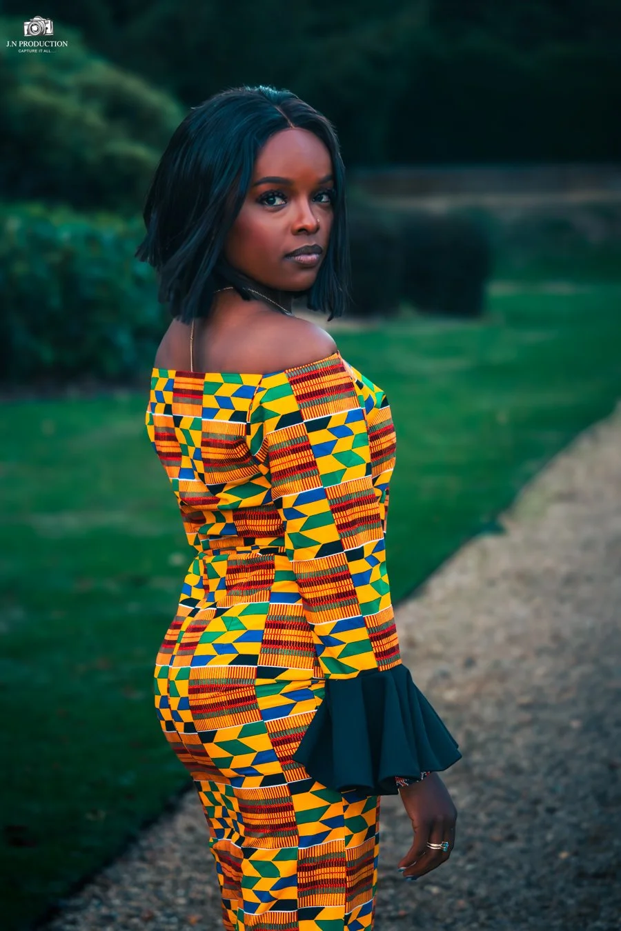A woman in a colorful, traditional African print dress features a pattern of yellow, green, blue, and red geometric shapes. She standing outdoors on a gravel path with greenery in the background, looking over her shoulder.