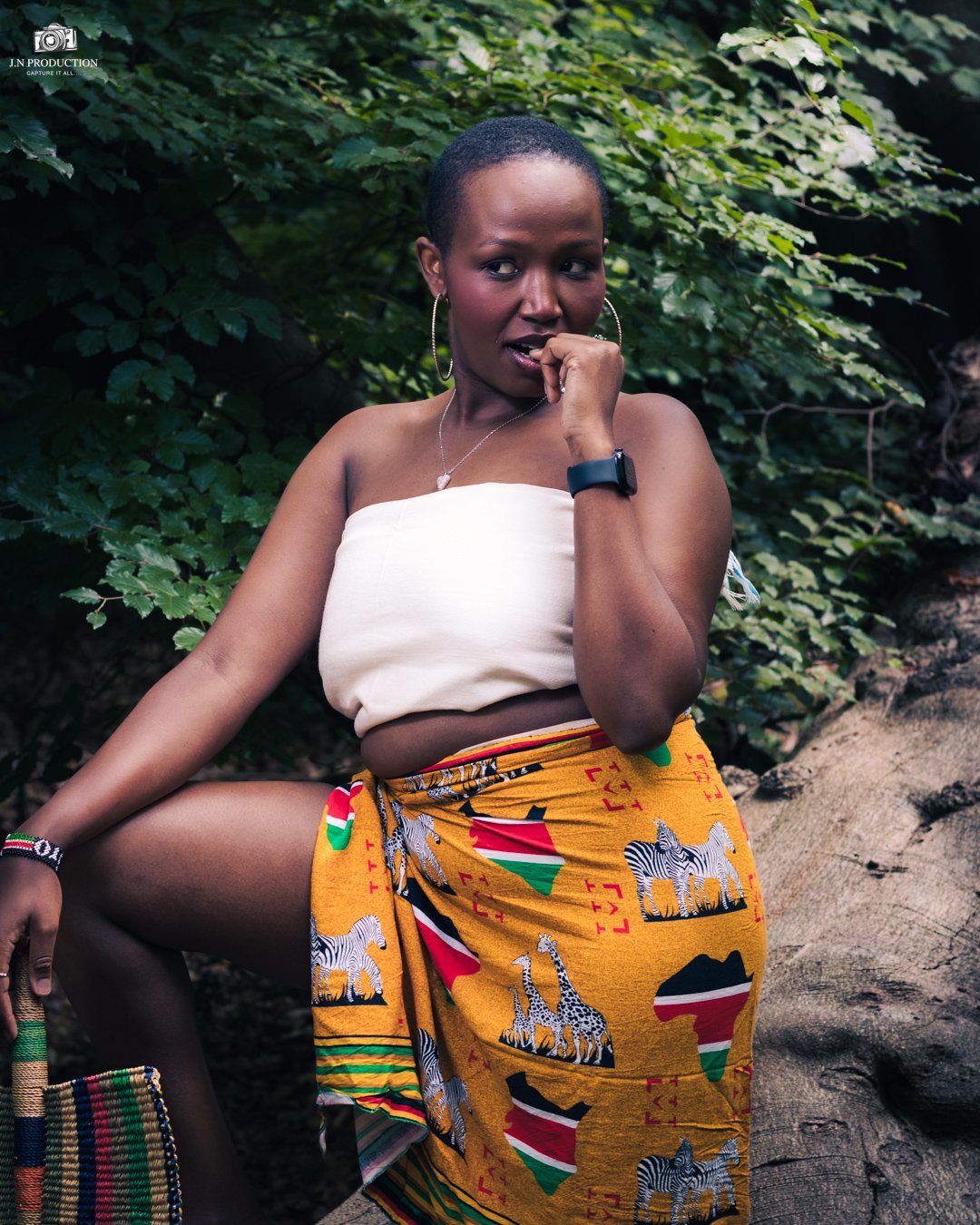 A woman with short hair wearing a white strapless top, colorful African print wrap with zebras and flags, accessorized with hoop earrings, a necklace, a smartwatch, and a bracelet, posing outdoors among green foliage.