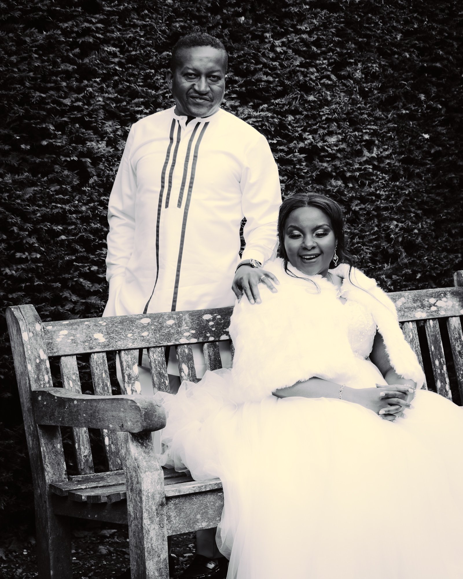 A man in a white traditional African outfit standing beside a woman in a wedding dress sitting on a wooden bench outdoors with a hedge background.