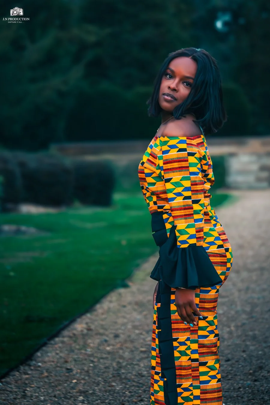 A woman with dark, shoulder-length hair standing outdoors on a gravel path, wearing a colorful, patterned dress with black accents, looking at the camera with a serious expression.