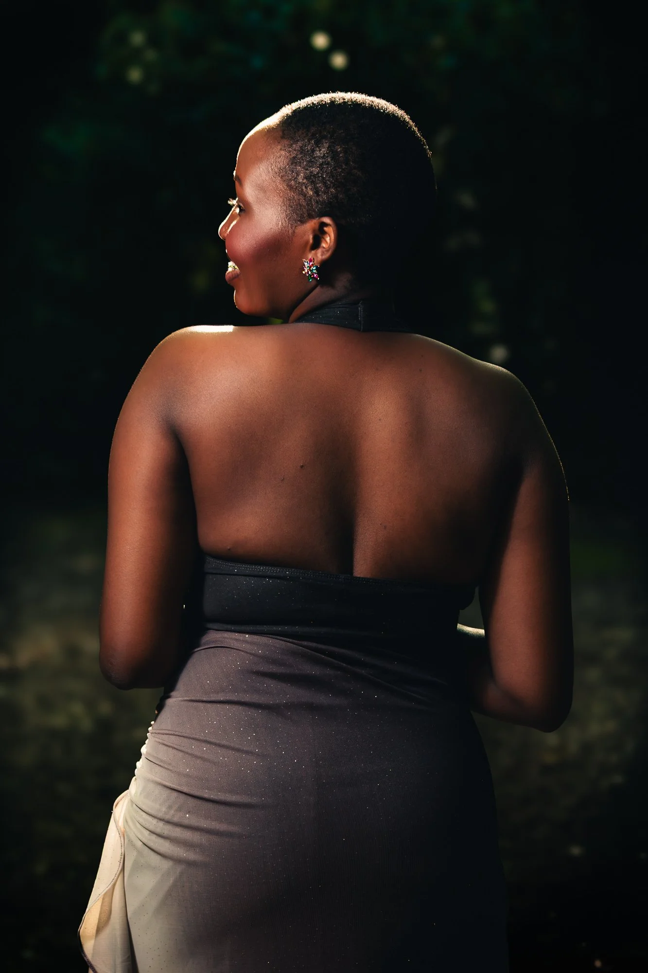 A woman with short hair and dark skin is standing outdoors at night, facing away from the camera, with her head turned slightly to the side. She is wearing a black strapless dress and colorful earrings, with soft lighting highlighting her shoulder an