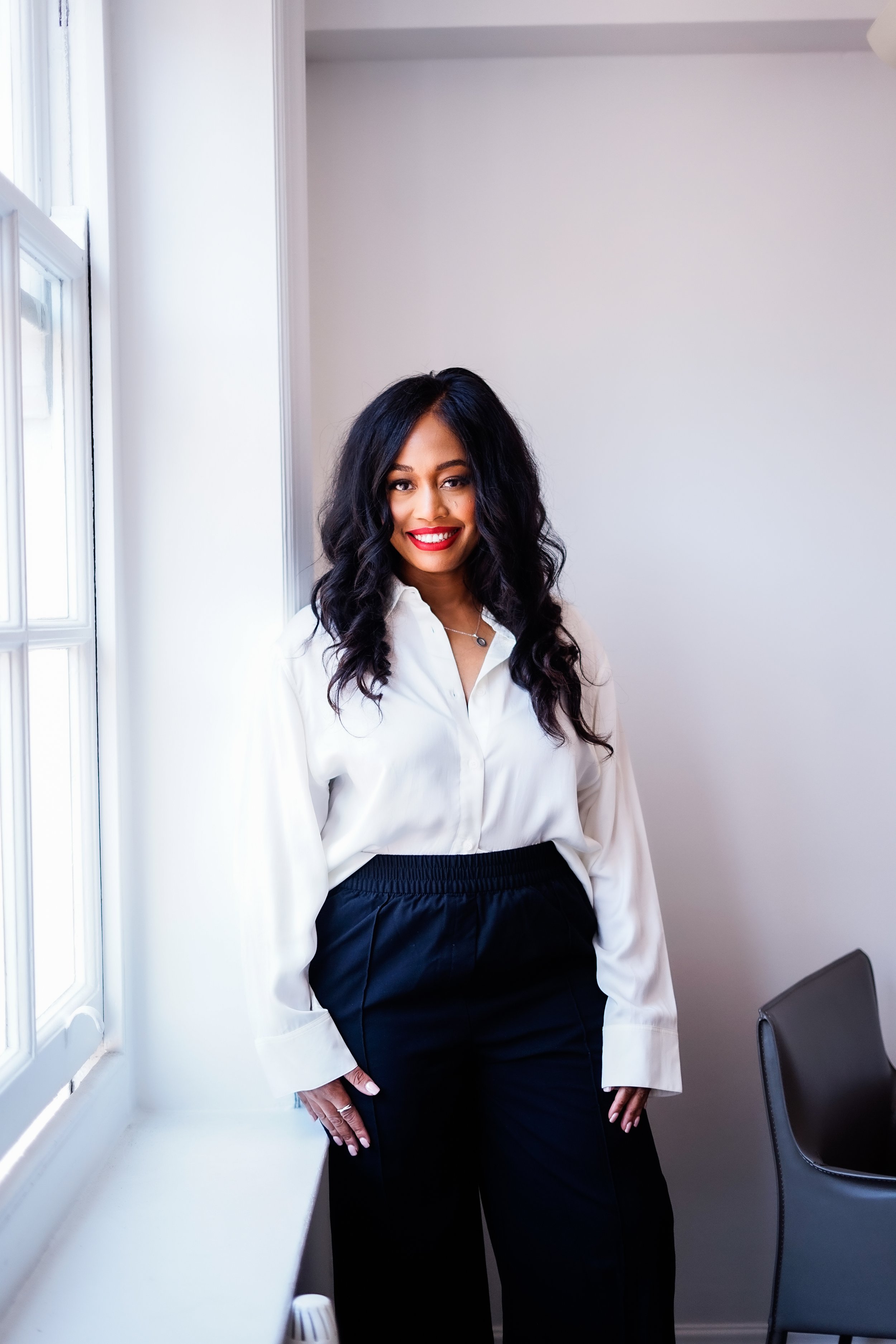 A woman with long, dark, wavy hair and red lipstick, wearing a white blouse and black pants, standing next to a window in a bright room.