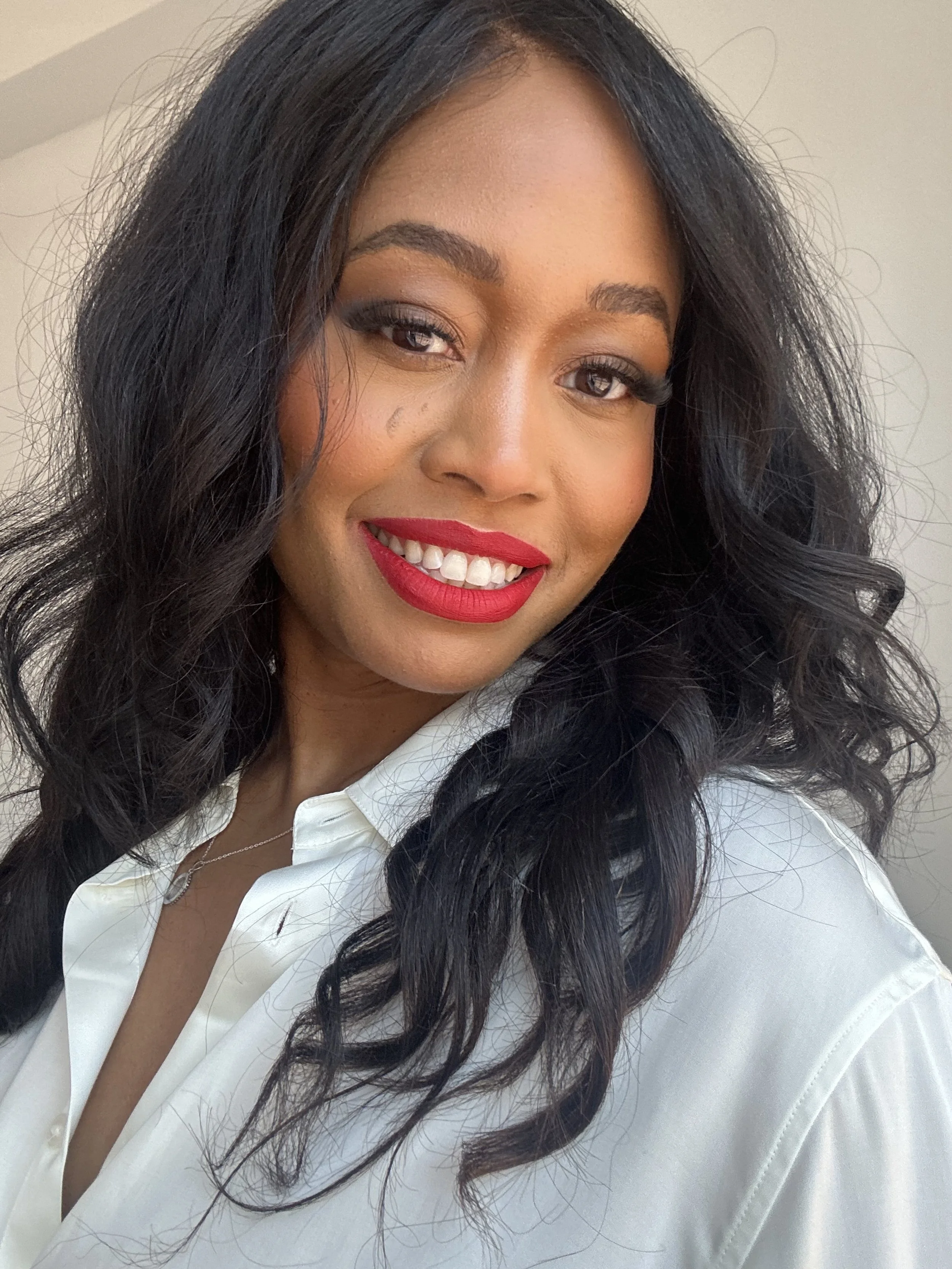 Close-up of a woman with long black curly hair, wearing a white shirt, red lipstick, and a silver necklace.