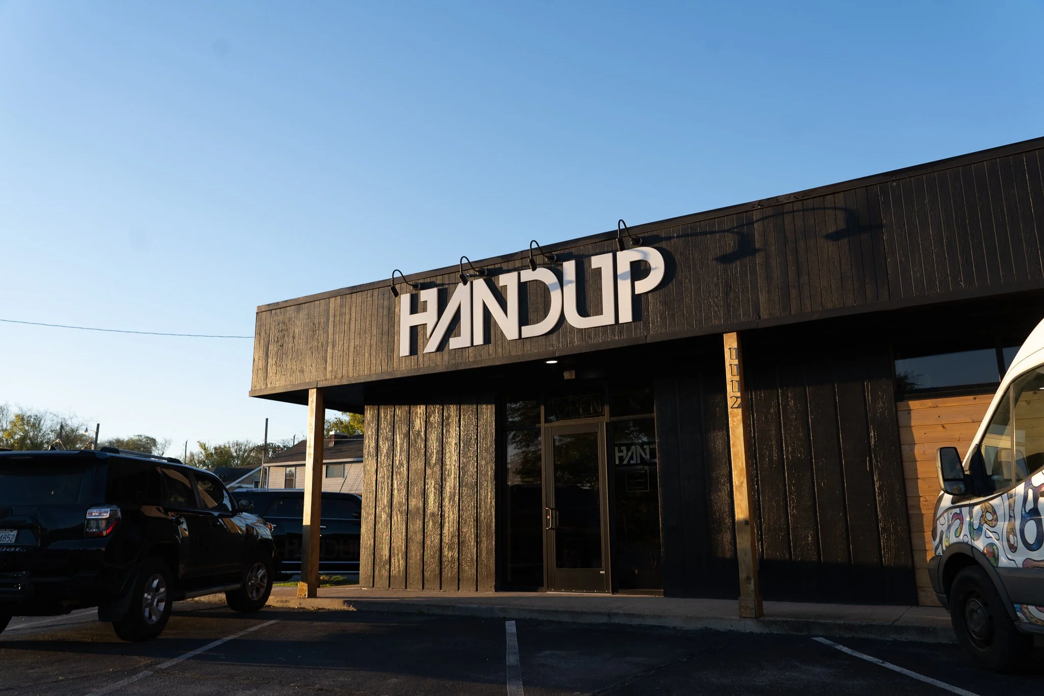 Exterior of a black wooden building with large white letters spelling 'HANDUP' on the sign, parking lot with cars in front, clear blue sky, and trees in the background.