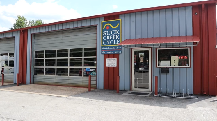Exterior of Suck Creek Cycle with a large garage door, a door with an 'OPEN' sign, and a bike rack.