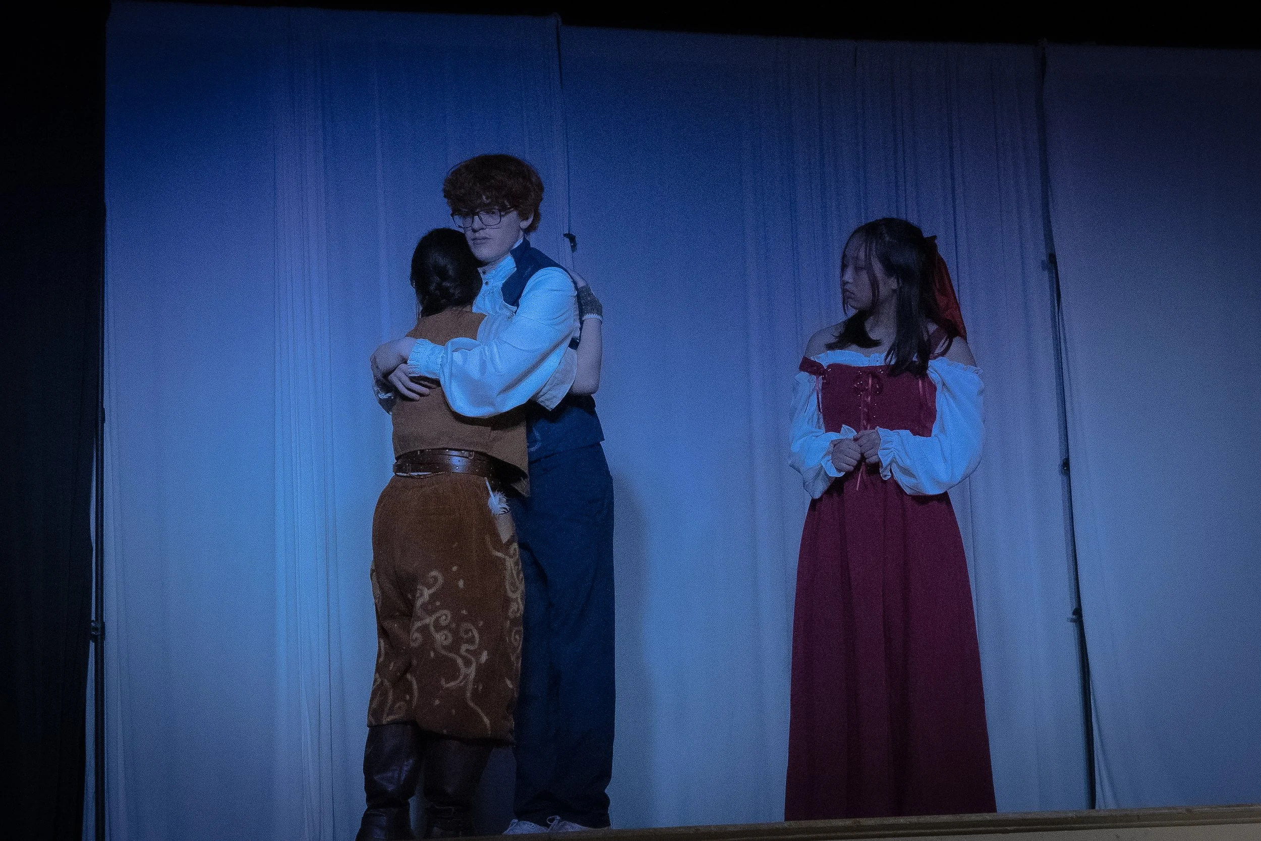 Three people on stage, with two embracing and one watching, in a theatrical setting with plain curtains in the background.