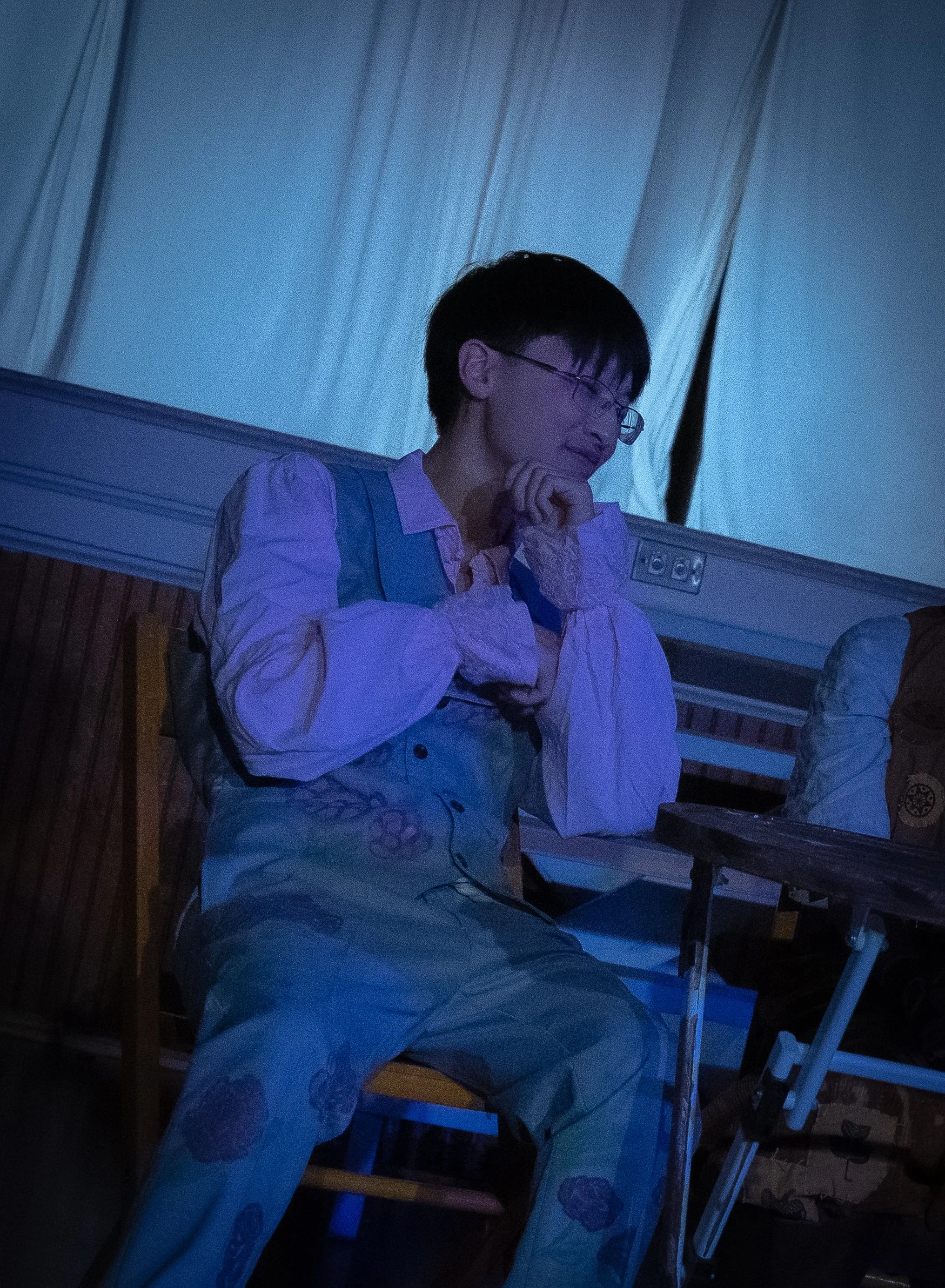 A person with short dark hair, glasses, and a patterned outfit sitting indoors in front of a light blue curtain, with a thoughtful pose and their hand near their chin.