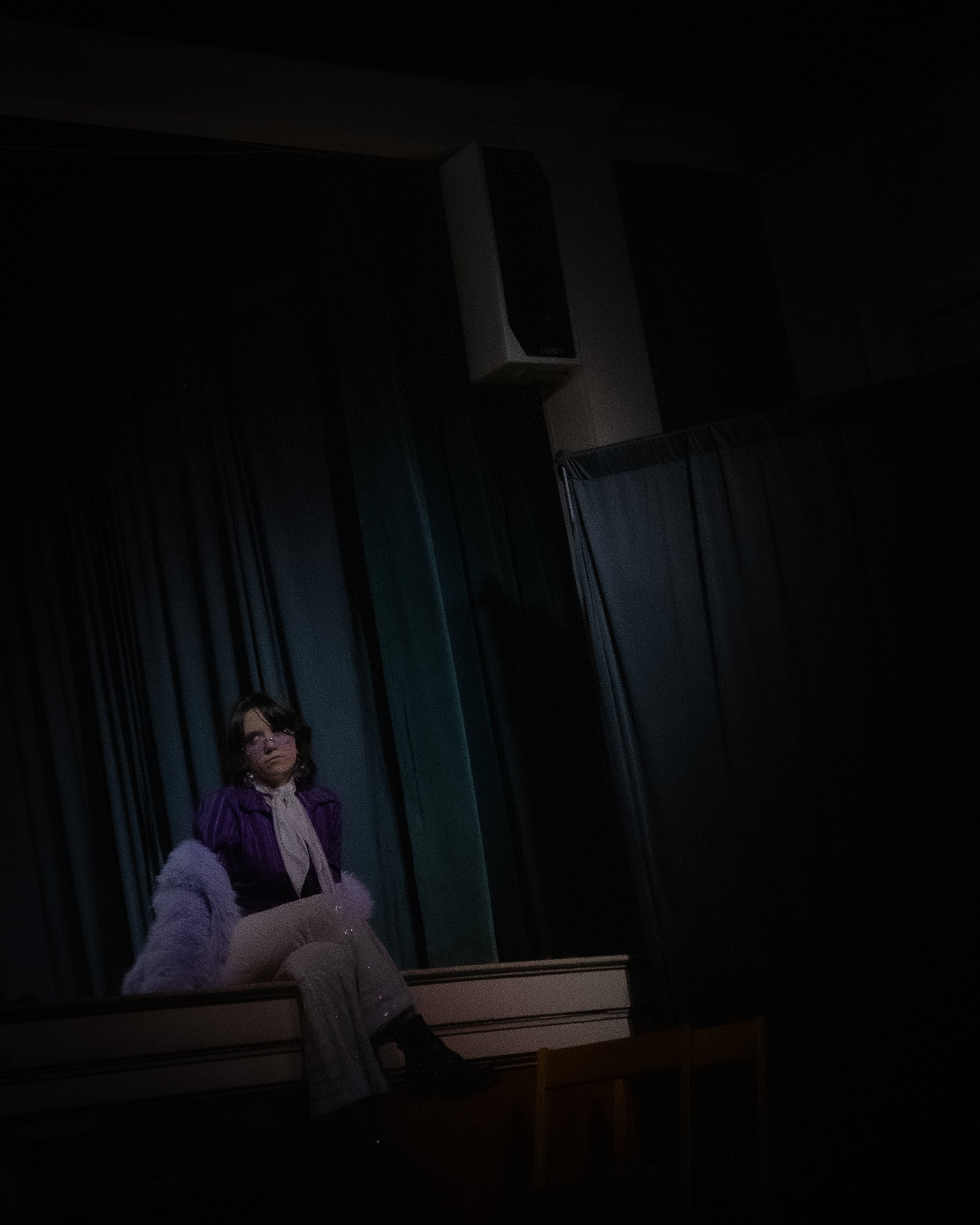 A woman sitting on a stage with green curtains, wearing a purple jacket, light-colored pants, black shoes, and pink glasses, under dim lighting.