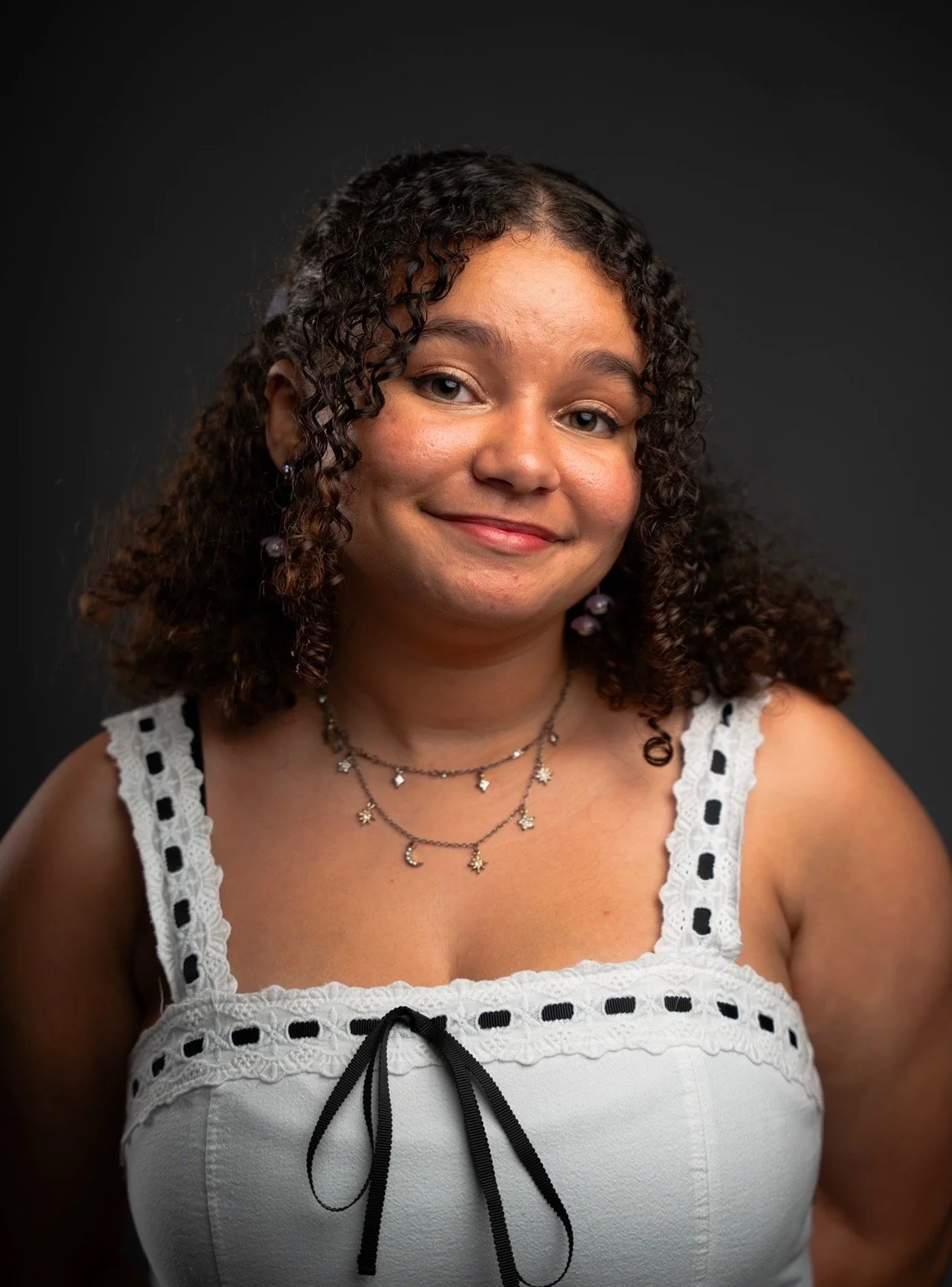 A young woman with curly hair, wearing a white sleeveless top with lace and black ribbon details, and decorative jewelry, smiling against a dark background.