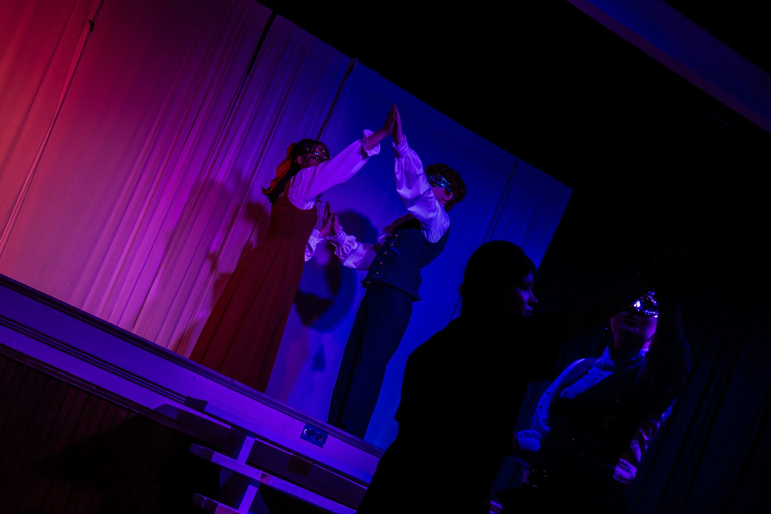 Two performers in costume with masks on stage, performing a synchronized dance with their hands pressed together, set against colorful stage lighting.