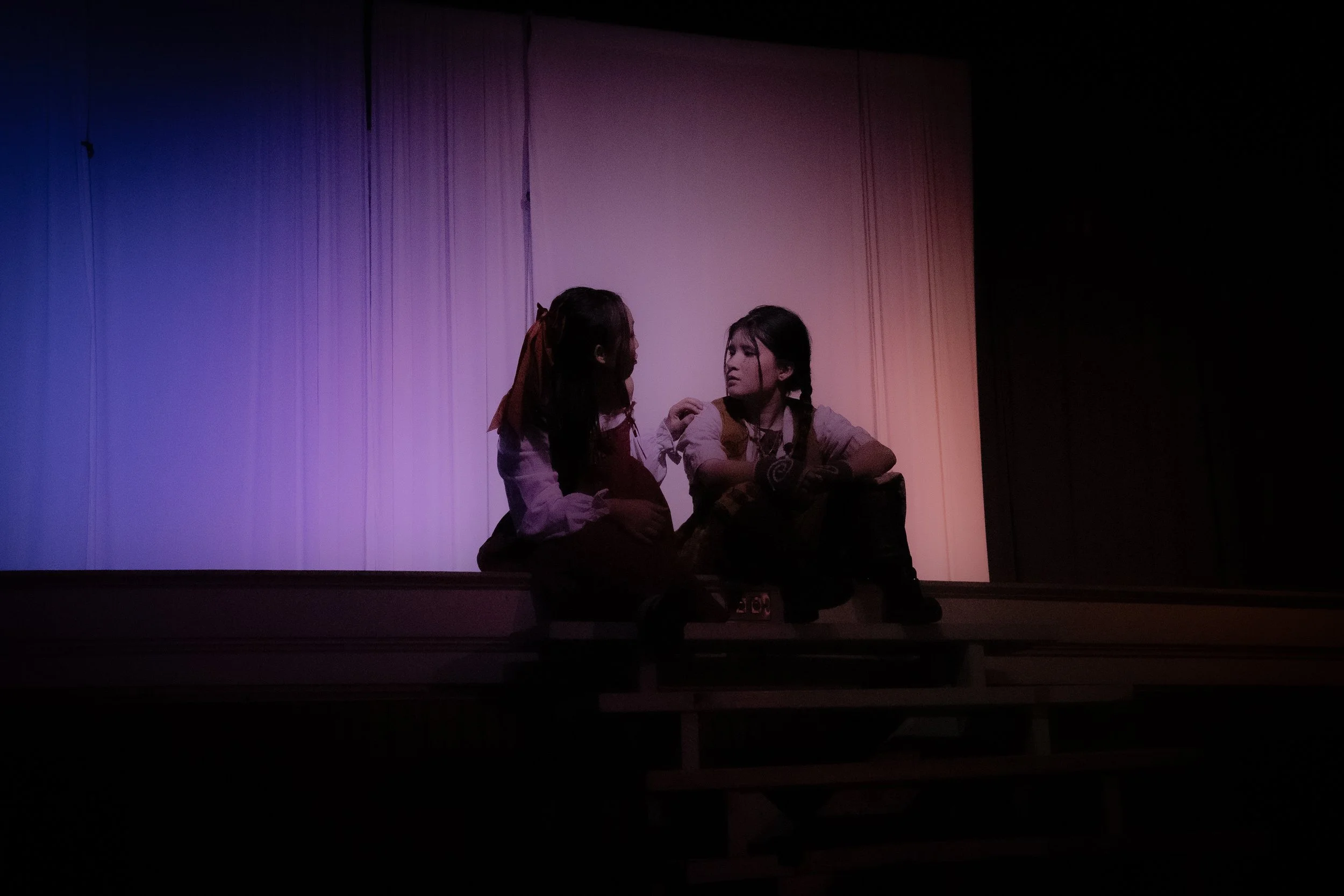 Two young women sitting on a stage with a colorful, softly lit curtain backdrop, engaged in conversation.