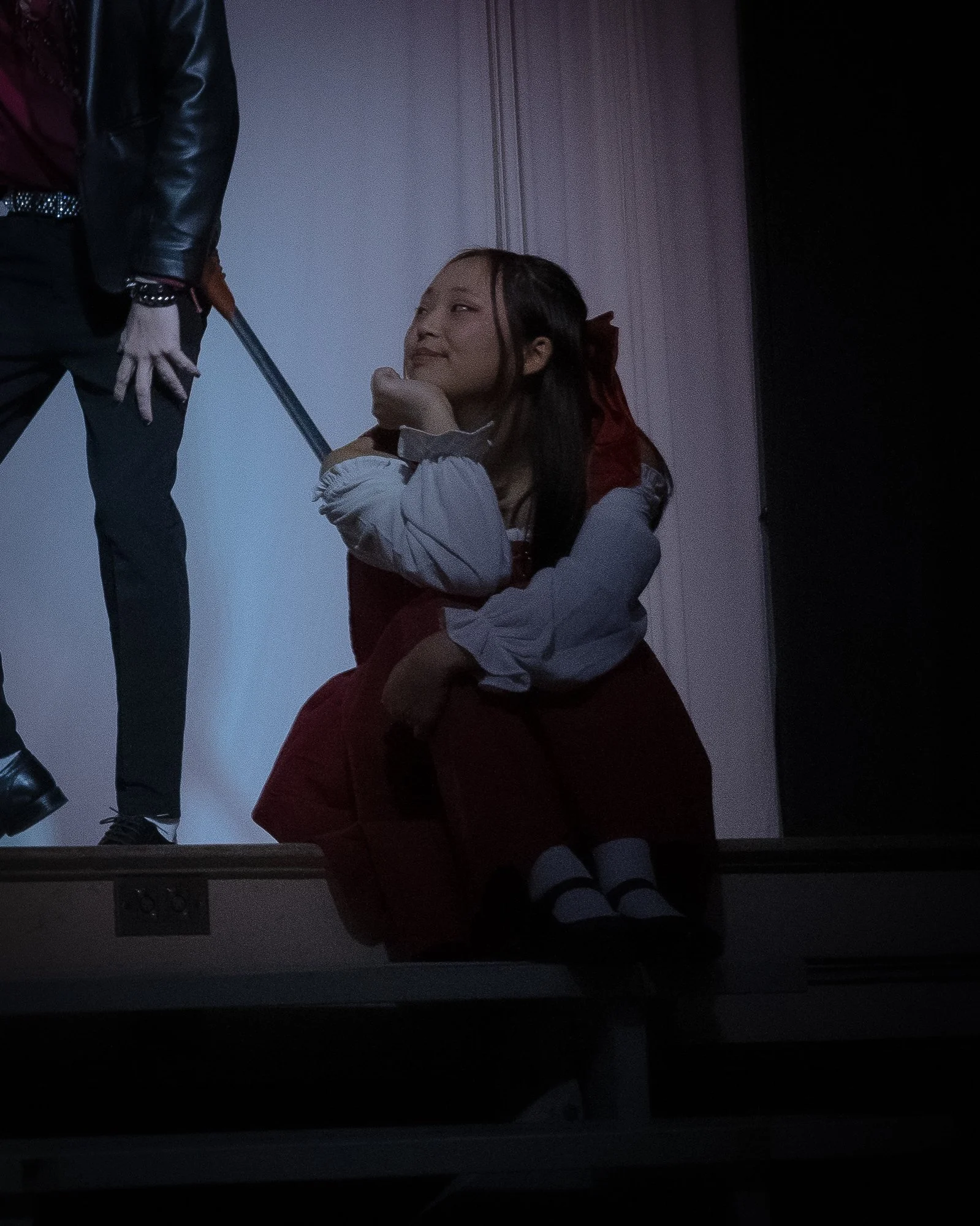 A girl with long dark hair, dressed in a red hooded cloak and white blouse, sitting on a stage with a person standing next to her holding a sword. The girl is smiling and resting her chin on her hand.
