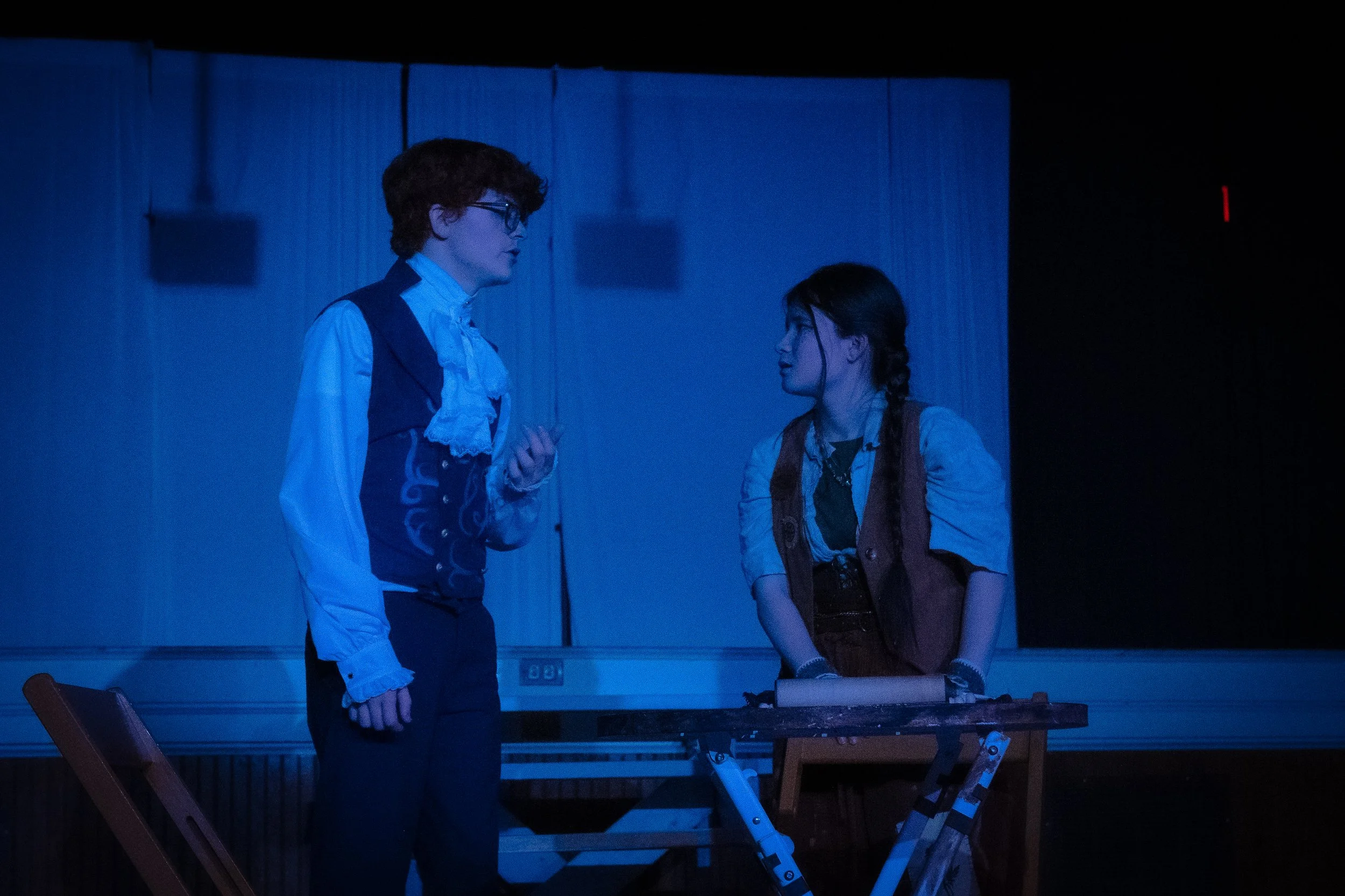 Two people on stage, one man and one woman, engaged in a conversation, with a blue-lit background and a table in front of them.