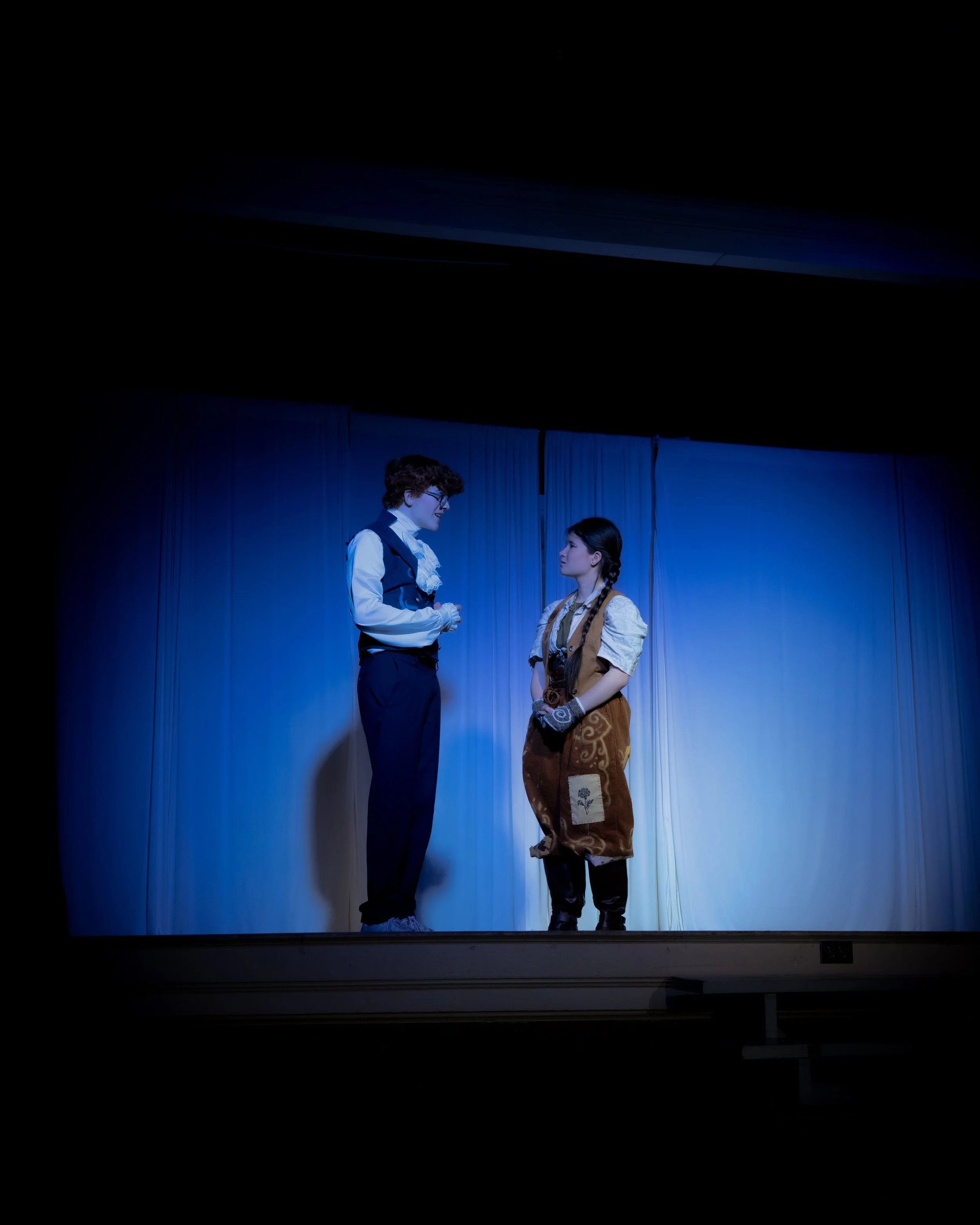 Two actors performing on stage, one dressed in vintage formal attire and the other in a costume resembling a traveler's outfit, with a blue curtain in the background.