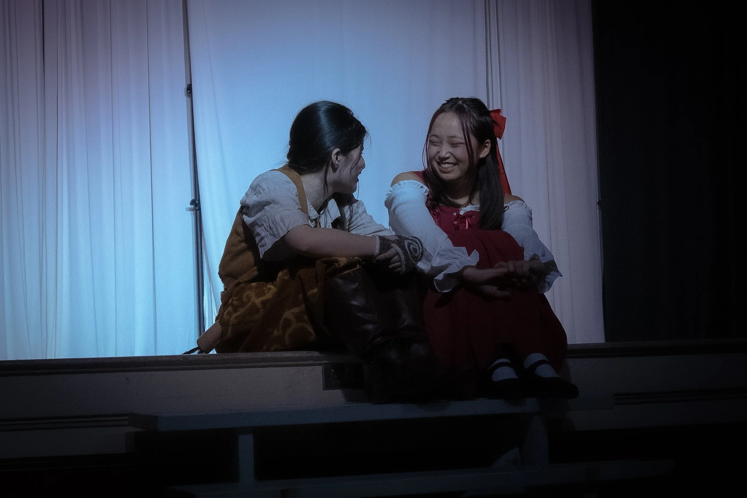 Two young women in costume on stage, sharing a moment of laughter. One is dressed as a girl with a red bow, the other in a vintage-inspired outfit, sitting on the stage with a curtain backdrop.
