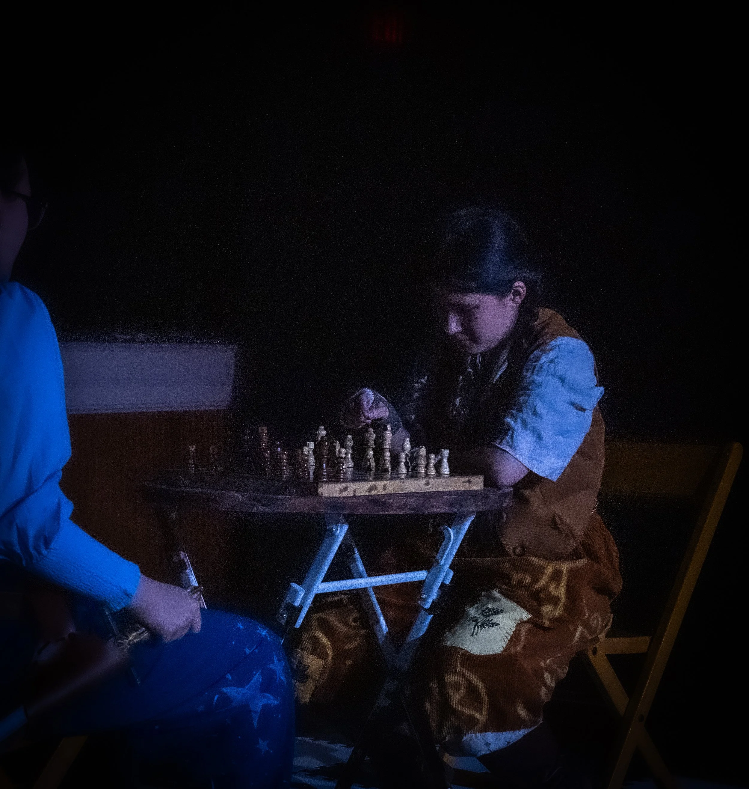 A young girl is playing chess in a dimly lit room, sitting at a small foldable table, with only her face and upper body illuminated.