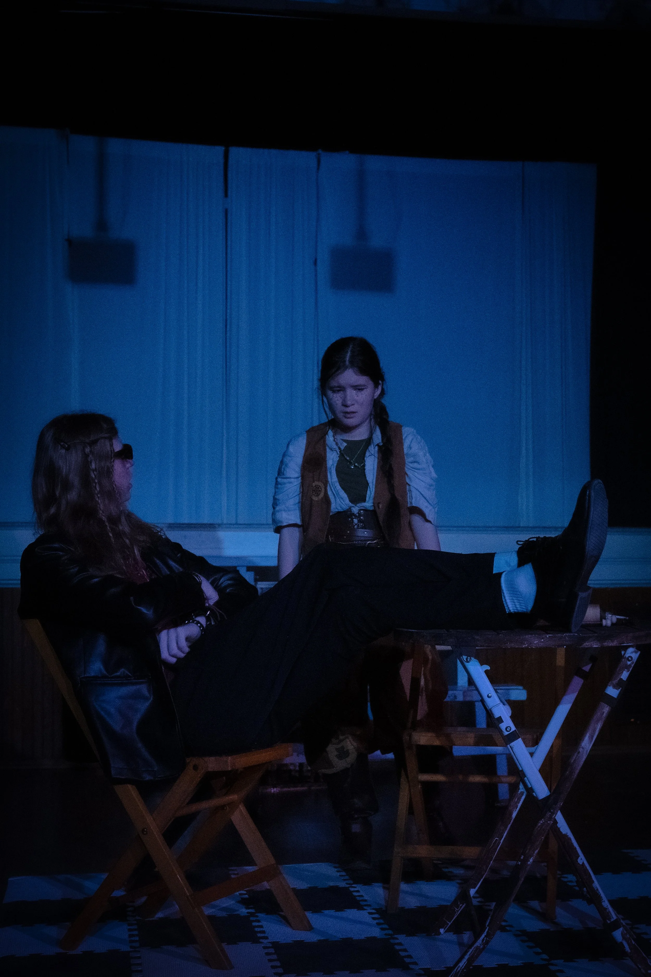Two young women on a stage, one seated with legs crossed and arms folded, wearing sunglasses and a leather jacket, and the other standing, looking down at her, with a blue background and a checkered floor.