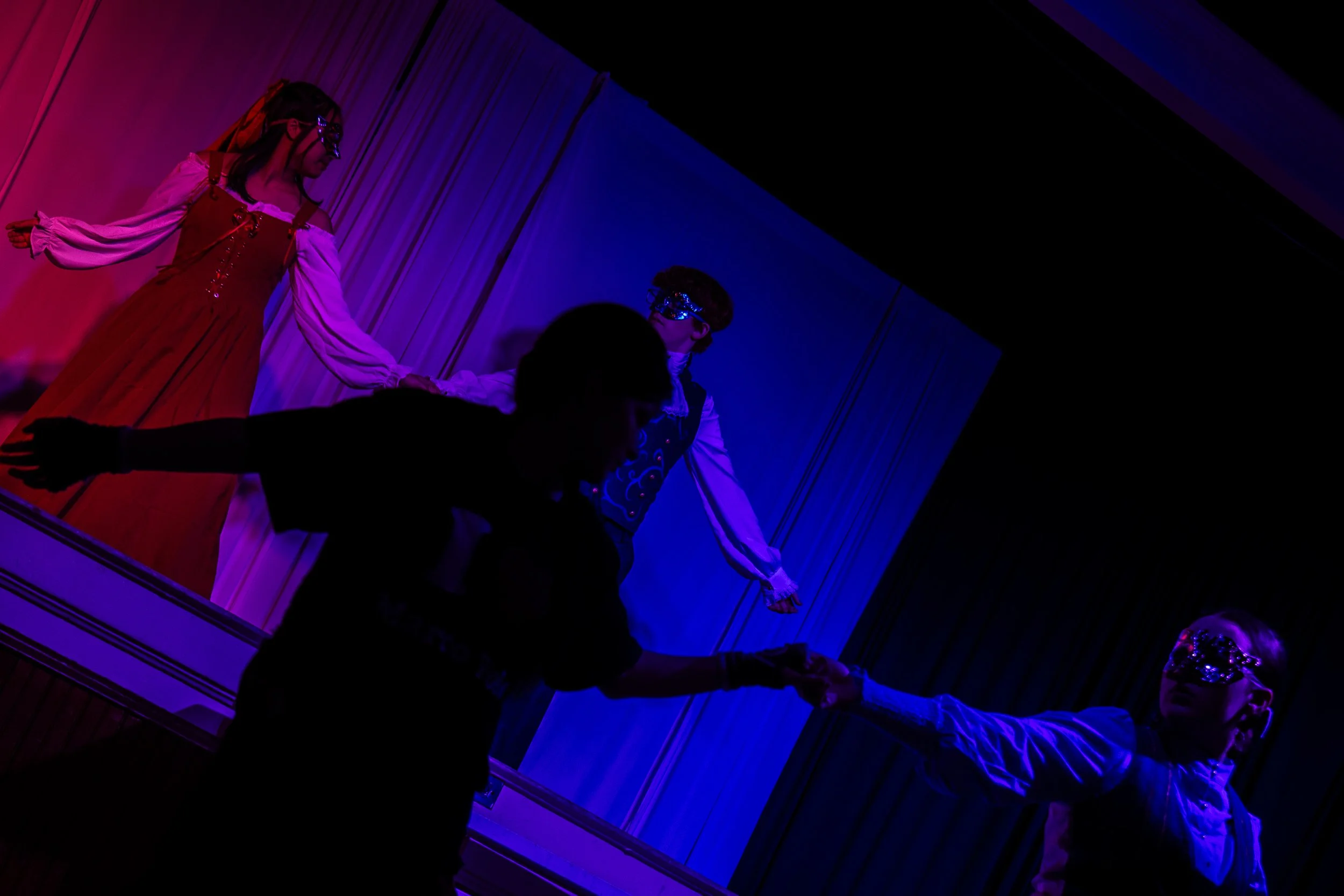 Two performers on stage wearing elaborate costumes and masks, holding hands, with dramatic colored lighting creating a theatrical atmosphere.