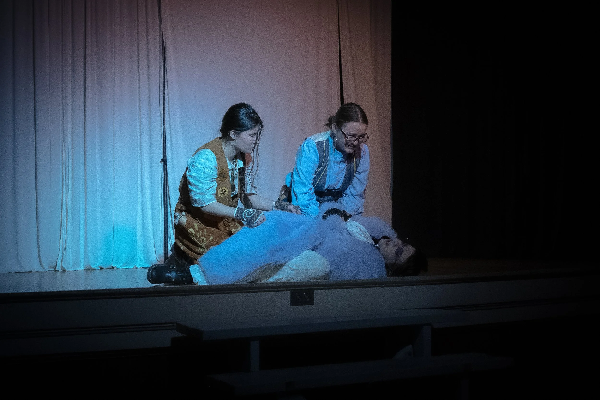 Two women in costumes appear to perform a scene on stage, with one lying down and the other two attending to her, in front of a plain curtain backdrop.