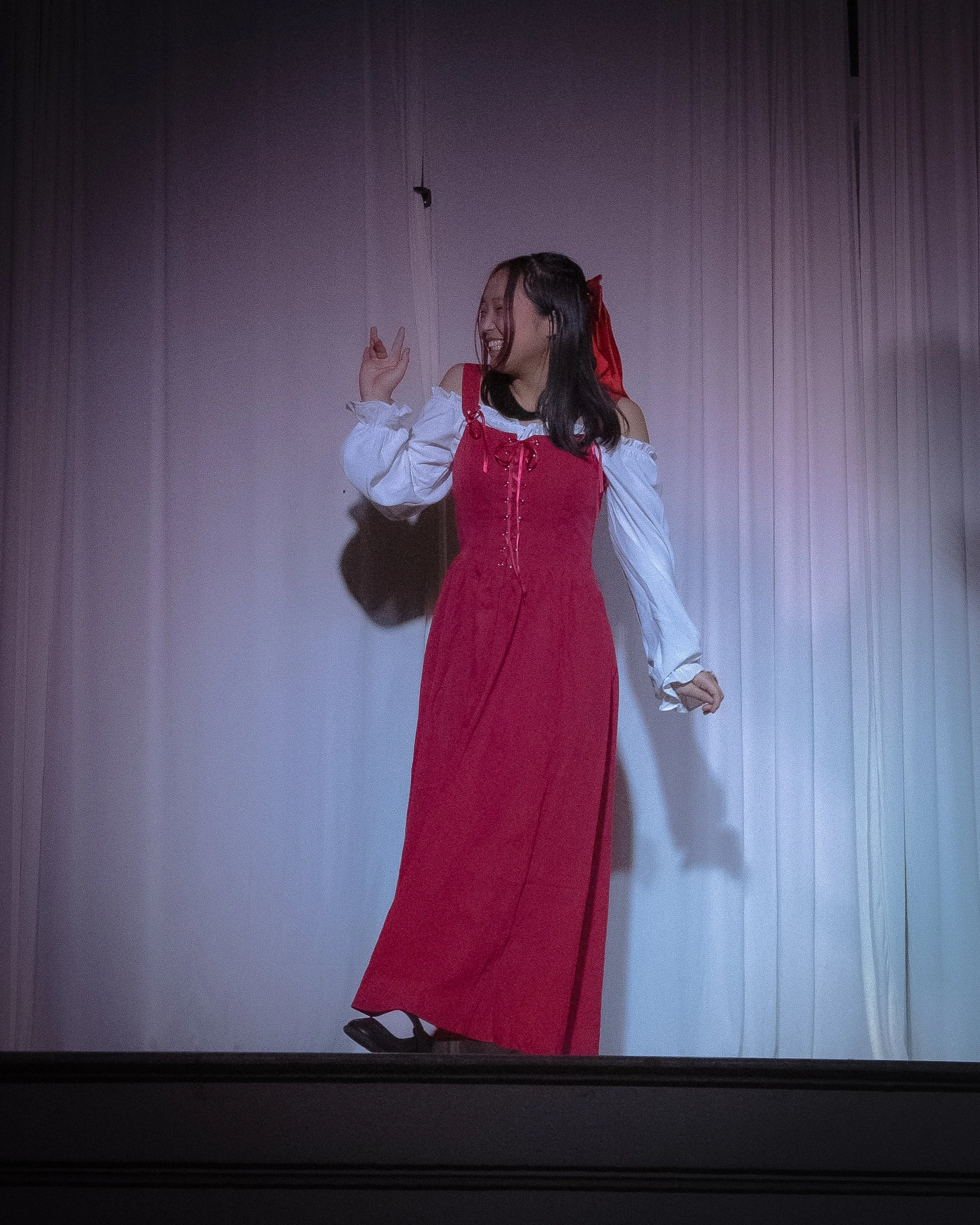 A woman dressed in a red and white costume on stage, smiling and gesturing with her hand.
