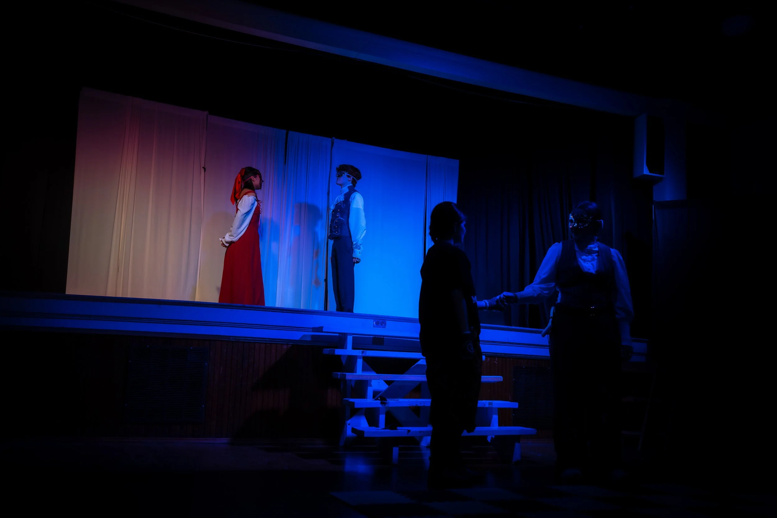 People performing on a stage with a curtain backdrop, two individuals on stage are facing each other, one dressed in a red dress and the other in a suit. Two others stand below, one facing away and one in glasses, holding hands.